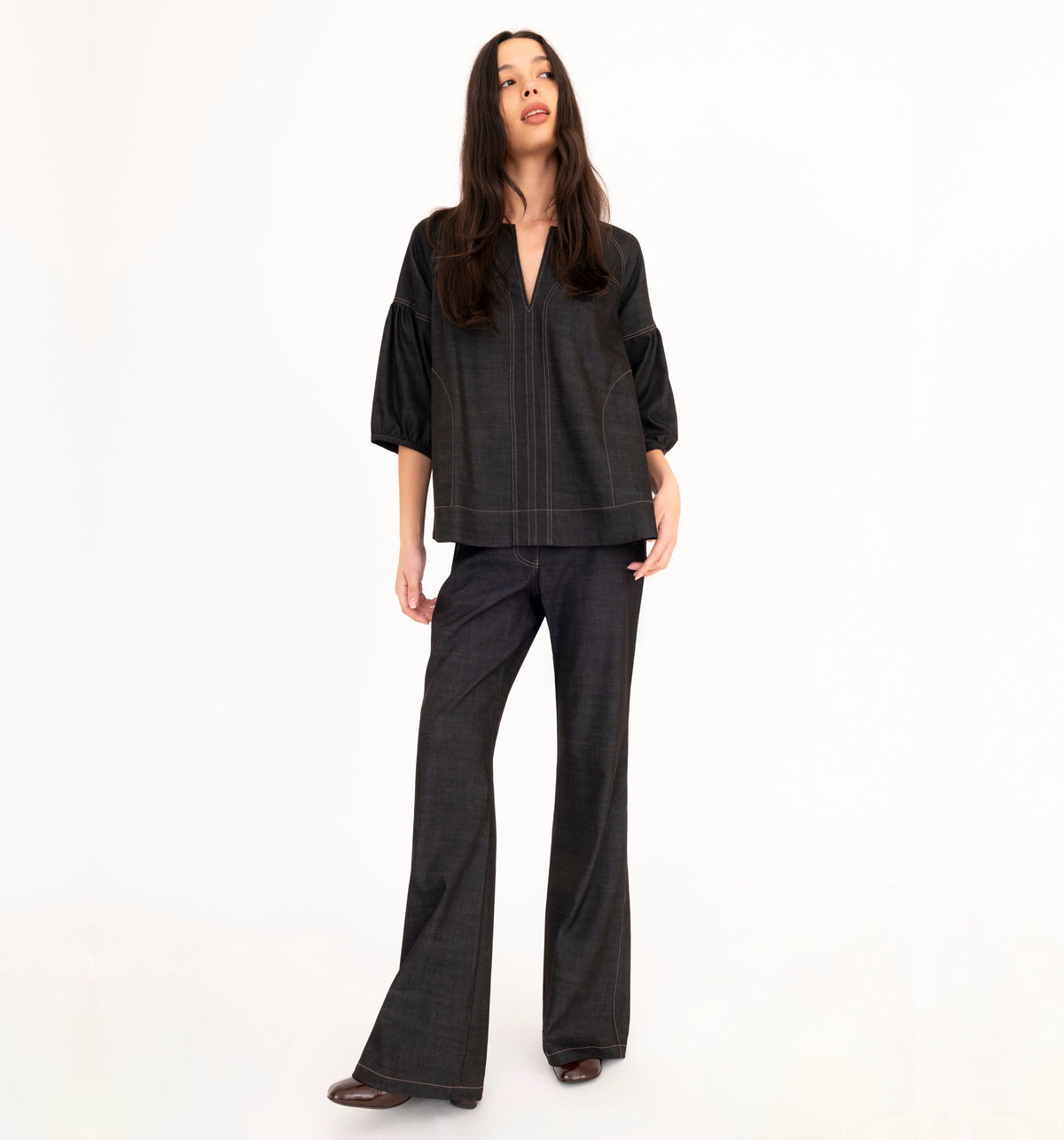 A woman modeling a black denim top with vertical seam details, paired with black denim flare pants and brown boots, against a plain white studio background.
