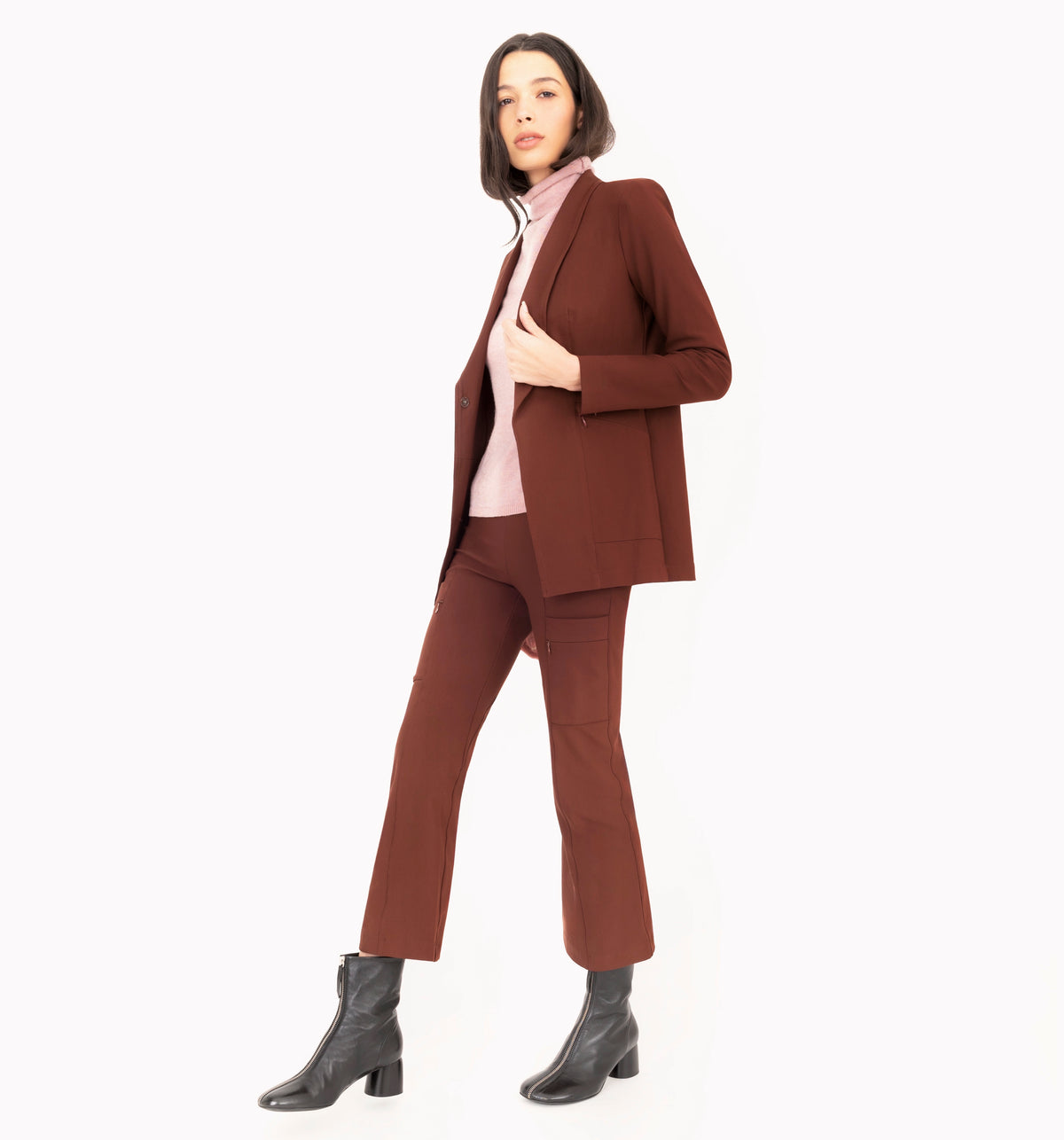 A model wearing a brown tailored pantsuit with a blazer and matching cropped pants, layered over a light pink cashmere turtleneck and paired with black ankle boots, shot against a solid white background.