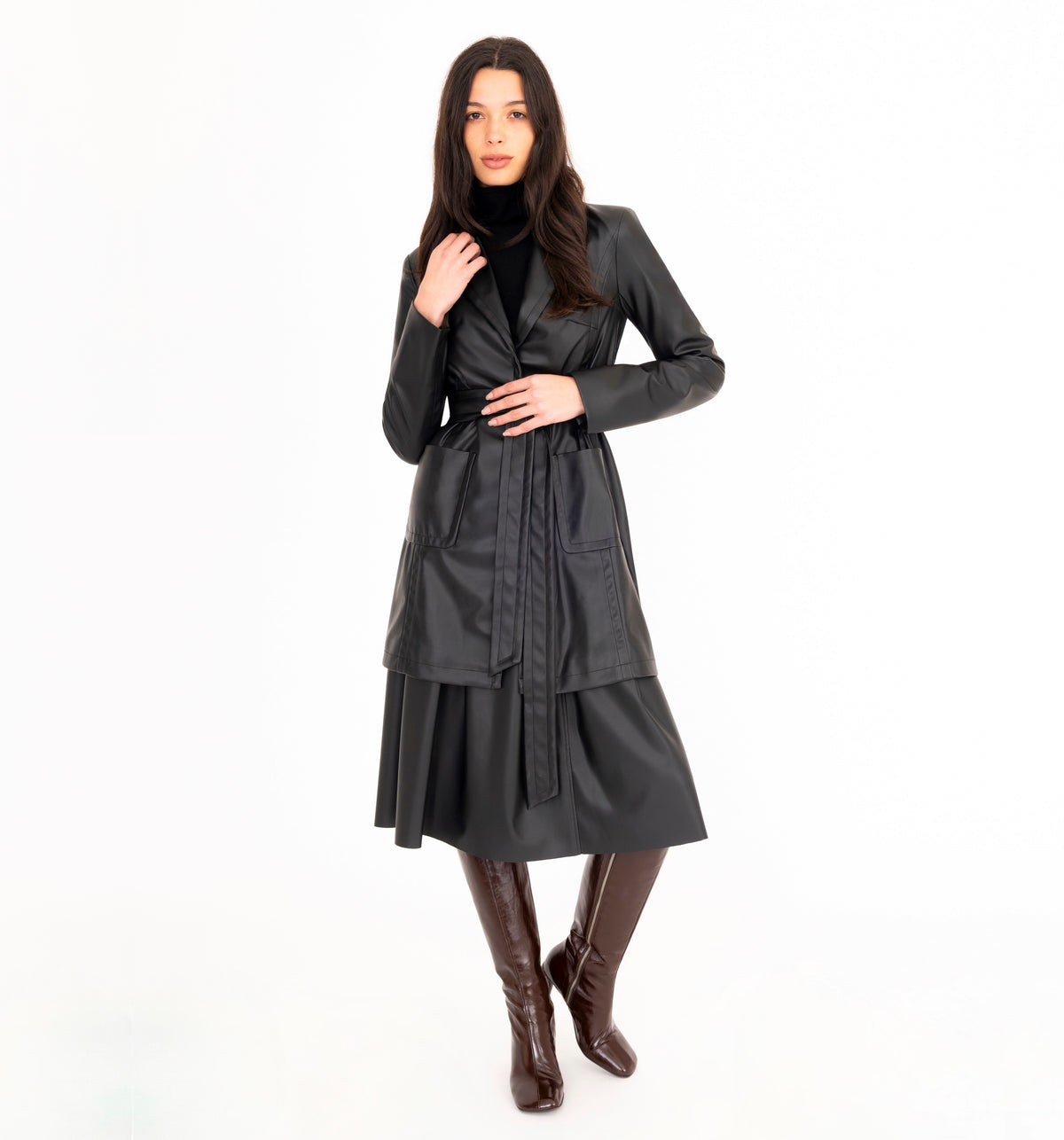 A woman modeling a black vegan leather trench coat and a pleated skirt, paired with brown knee-high heeled boots, against a plain white studio background.