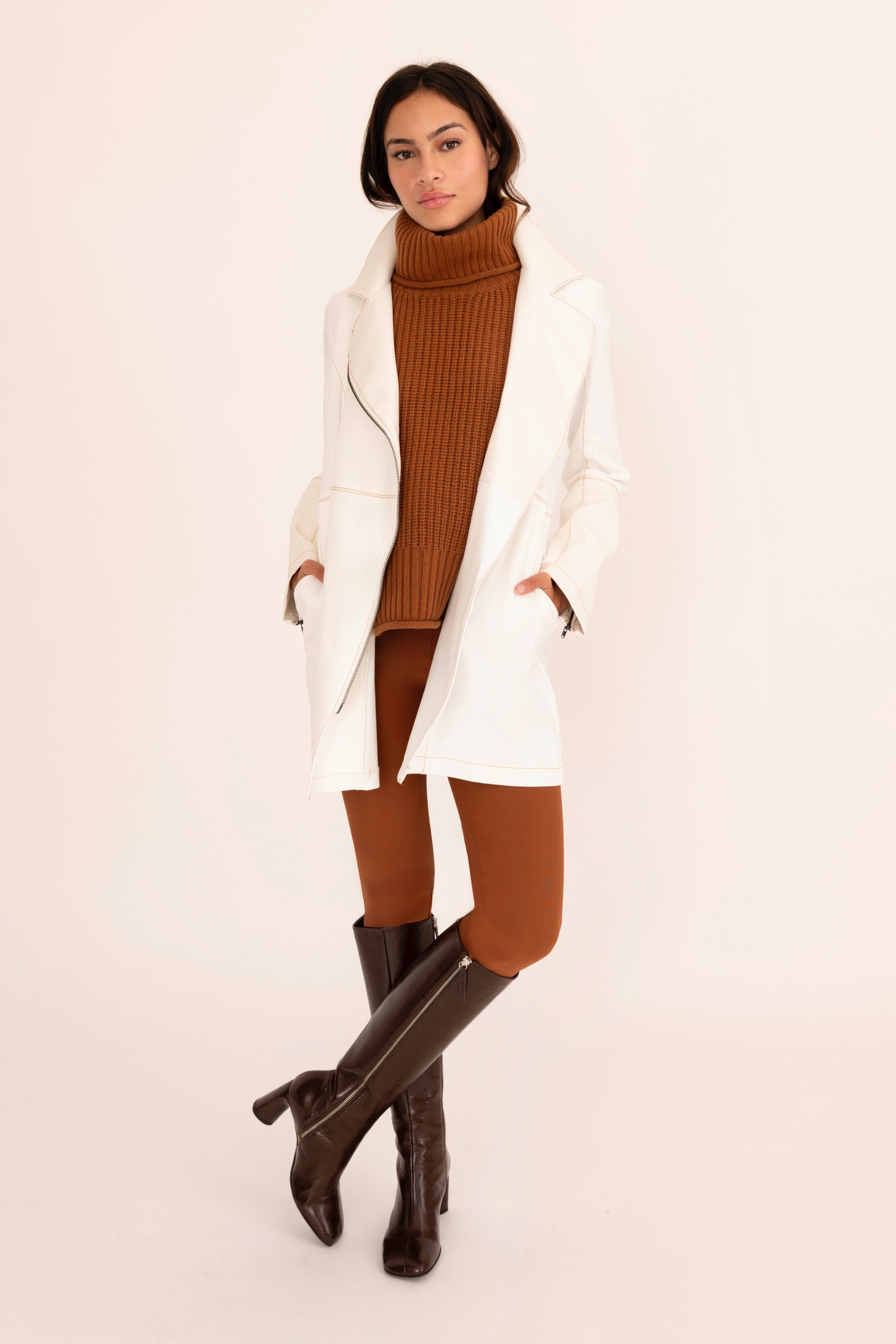 Woman wearing a white coat, brown turtleneck sweater, and knee-high brown boots on a beige background