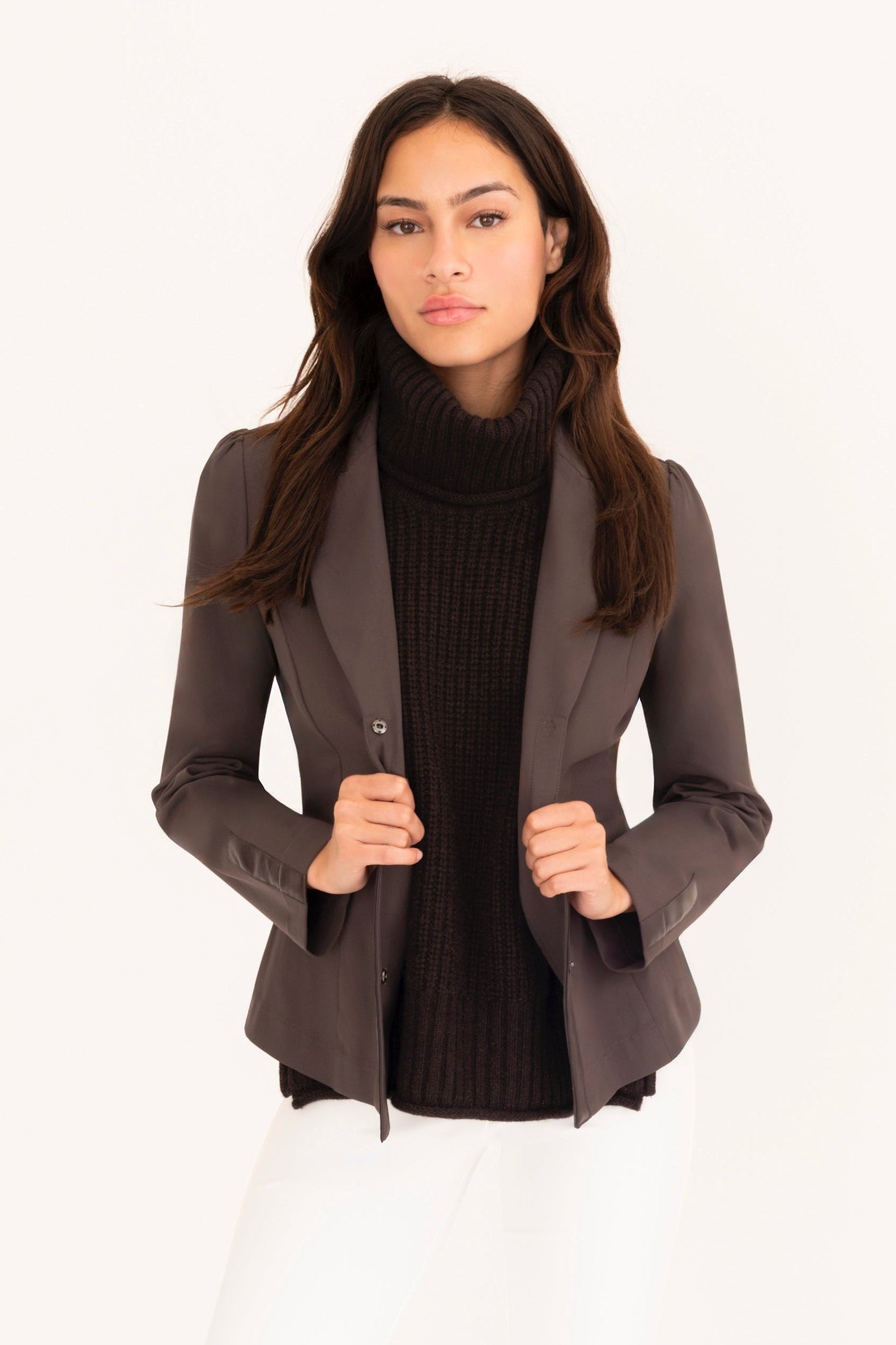 Woman wearing a brown blazer, brown top, and white pants on a white background