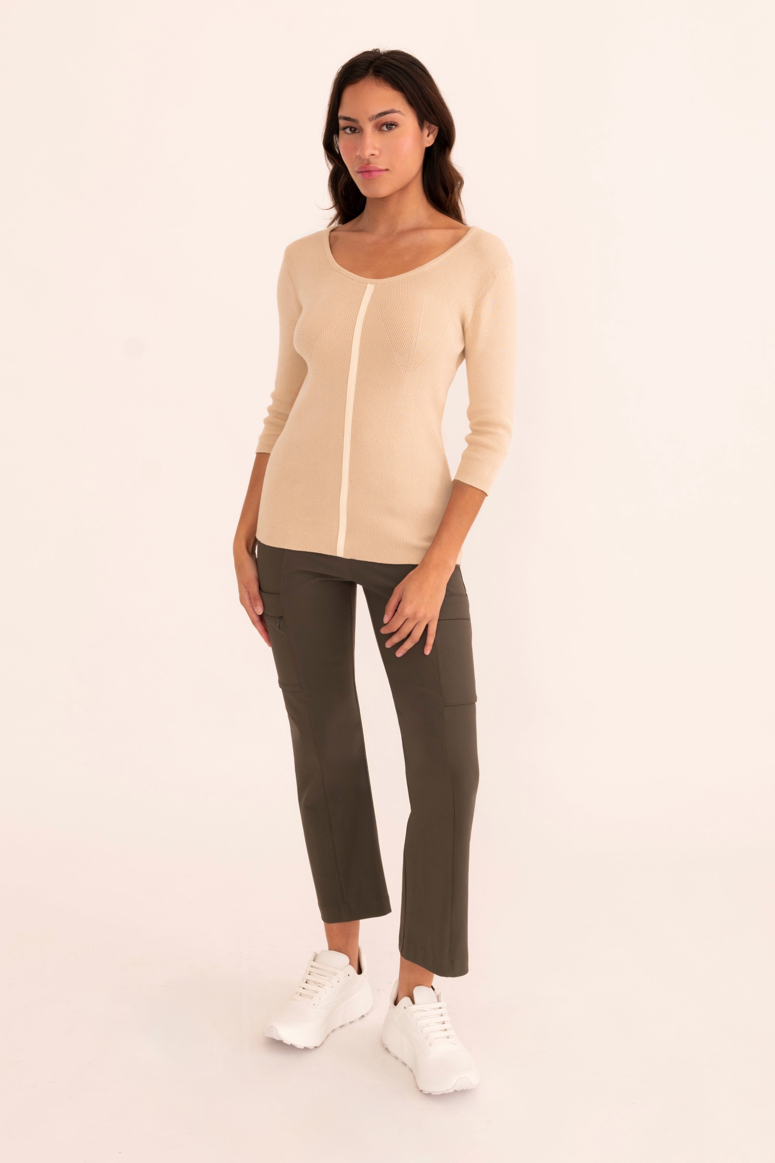 A woman modeling a beige three-quarter sleeve knit top paired with green cropped pants and white tennis shoes, standing against a plain white studio background.