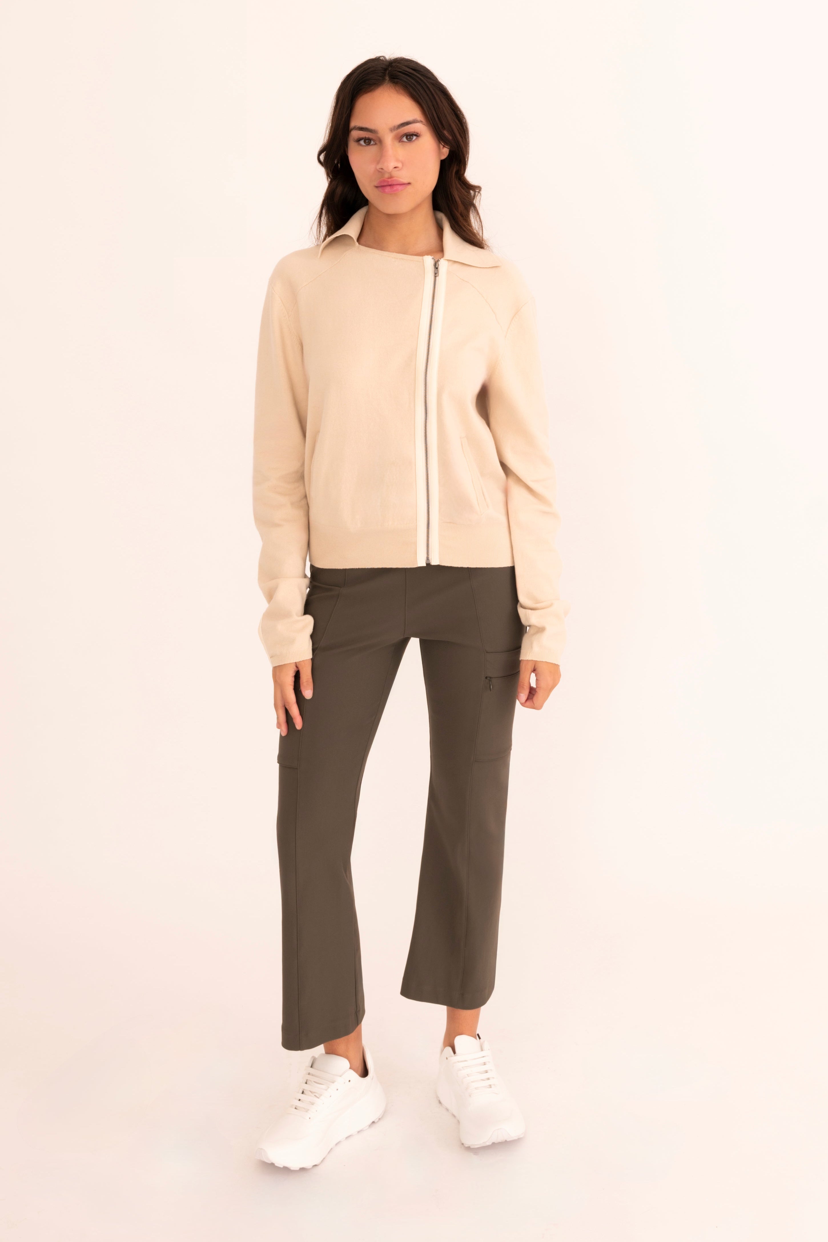 A woman modeling a beige zip-up knit cardigan with green cropped pants and burgundy loafers, standing against a plain white studio background.