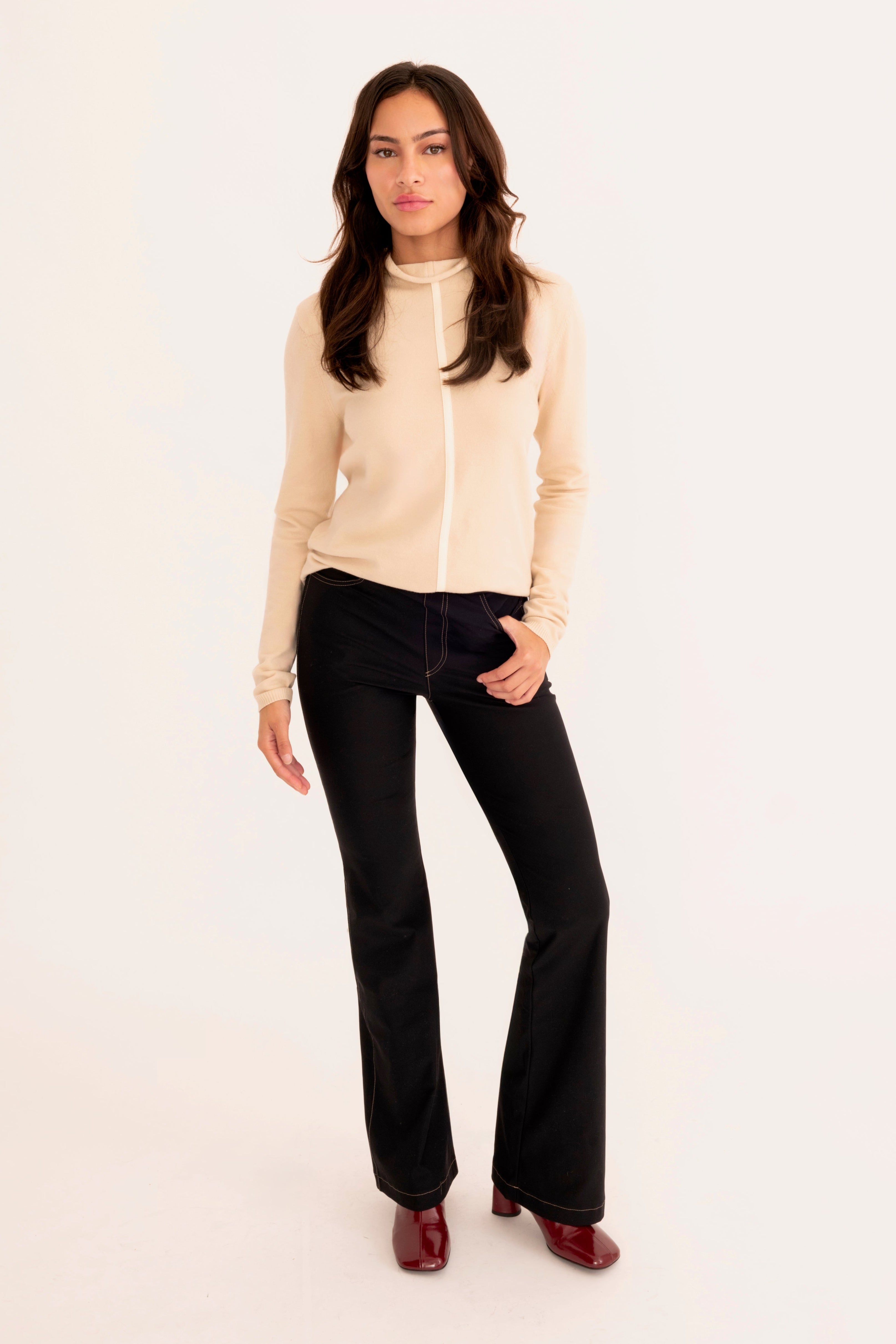 A woman modeling a beige mock-neck knit sweater and black flared pants against a plain white studio background.