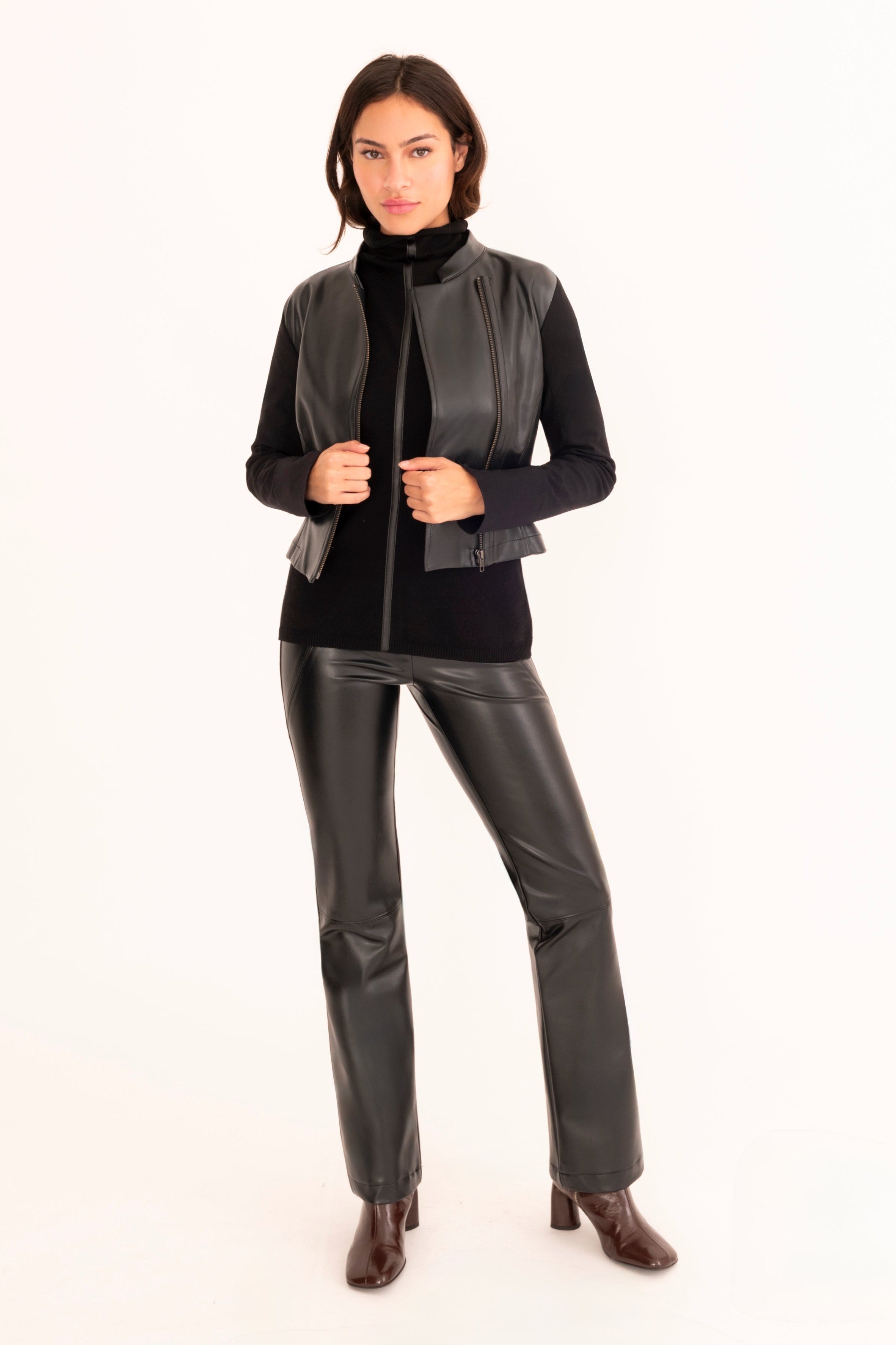 A woman modeling a black faux-leather jacket paired with matching pants standing against a plain white studio background.