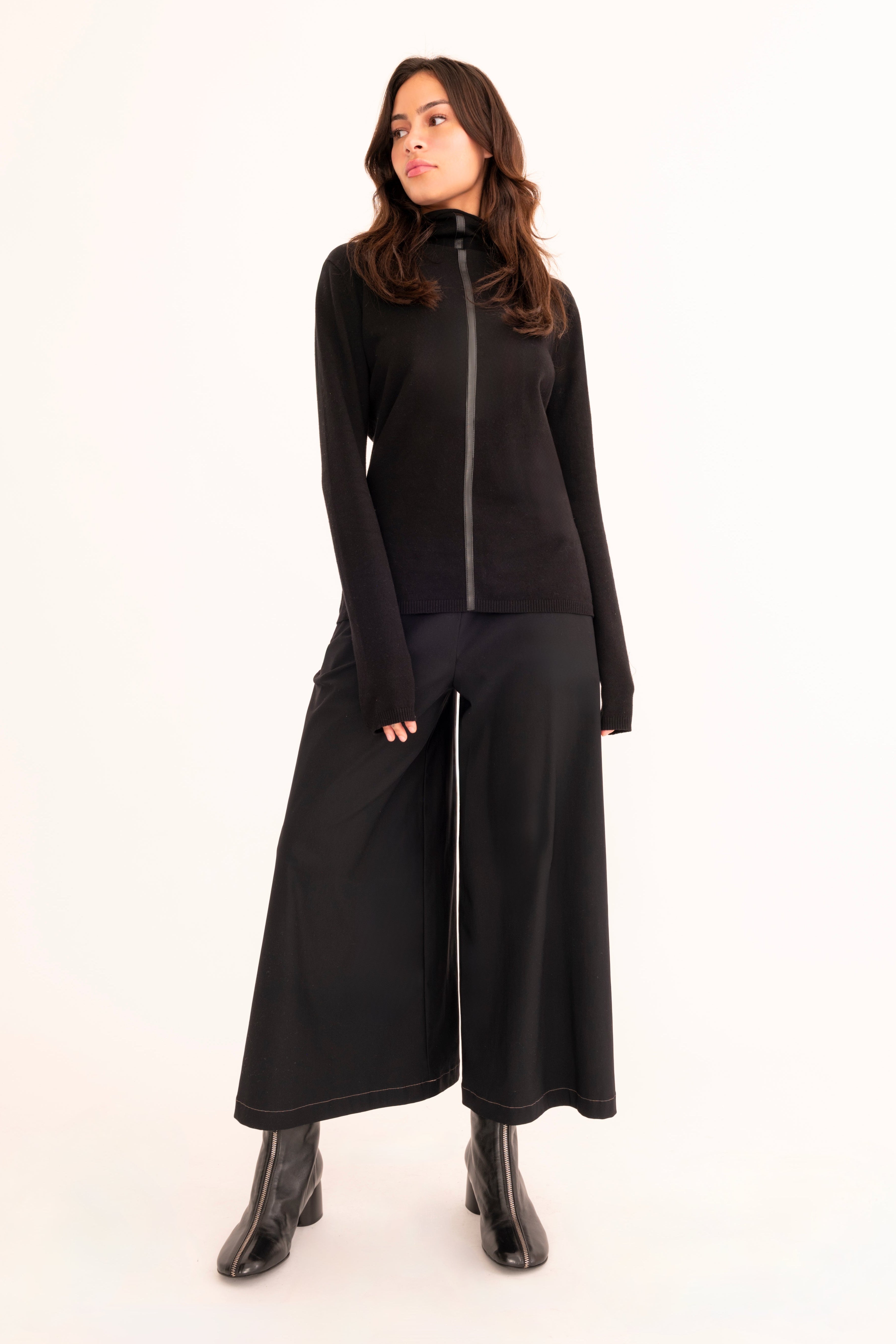 A woman modeling a pair of wide-leg cropped pants in black, paired with a black mock-neck long-sleeve top and black studded loafers, against a plain white studio background.