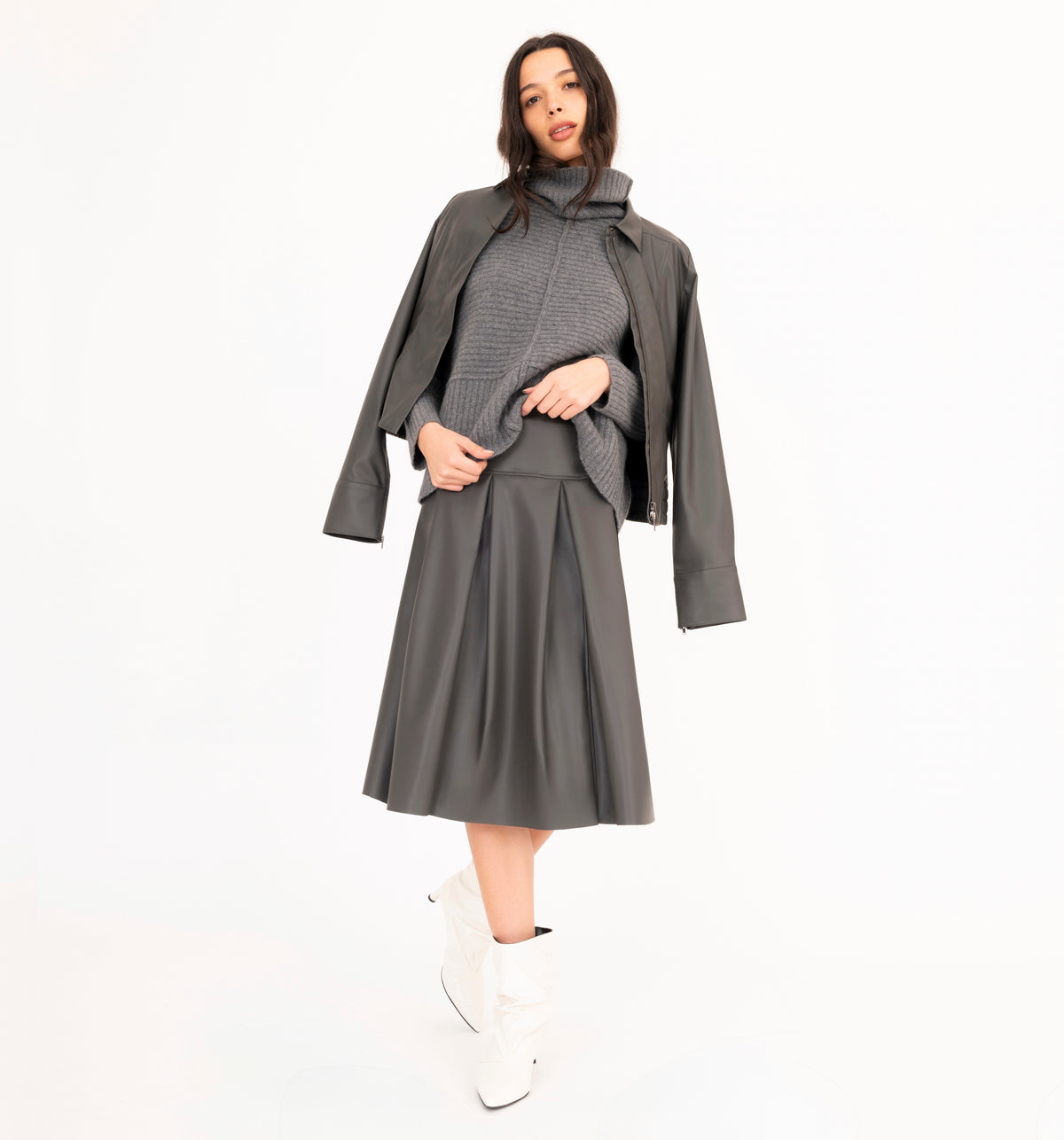 A woman wearing a gray vegan leather pleated skirt with a matching vegan leather jacket and cashmere funnel-neck sweater on a white background.