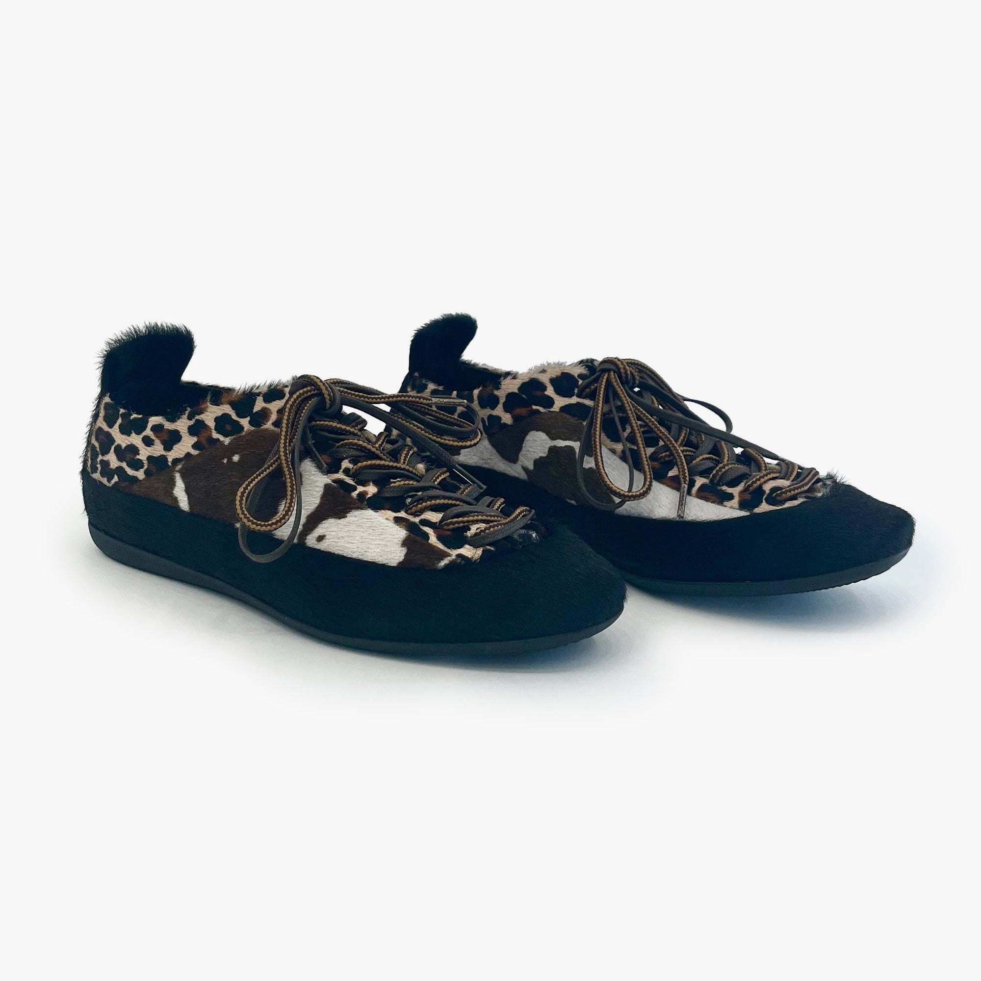 A pair of low-top slip-on shoes featuring a cow-print pattern on the sides, leopard print, black suede-like toe and heel sections, brown laces, and a black fur trim on the collar, shown against a plain light gray background.