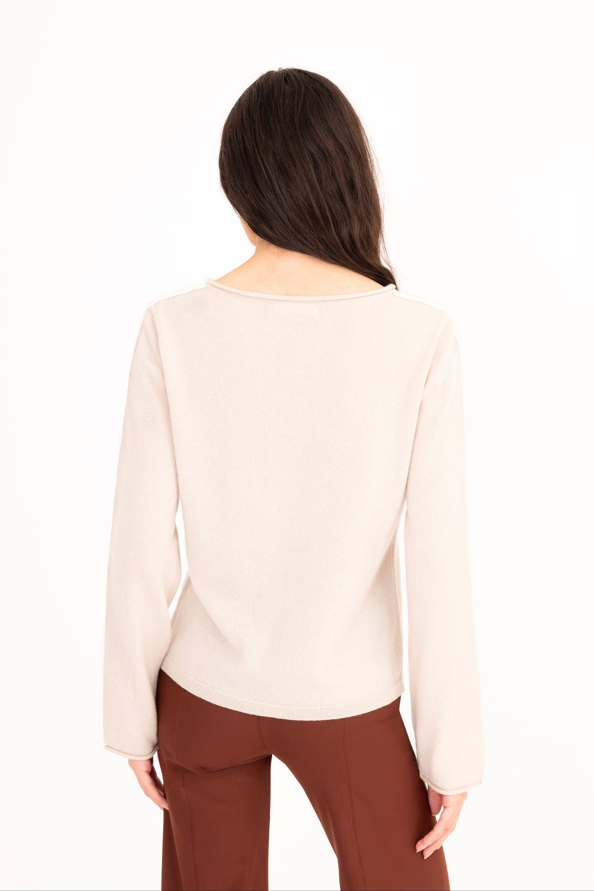 A woman modeling a beige long-sleeve knit sweater, paired with brown pants, against a plain white studio background.