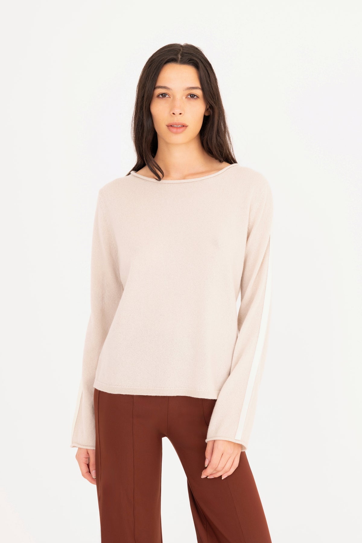 A woman modeling a beige long-sleeve knit sweater, paired with brown pants, against a plain white studio background.