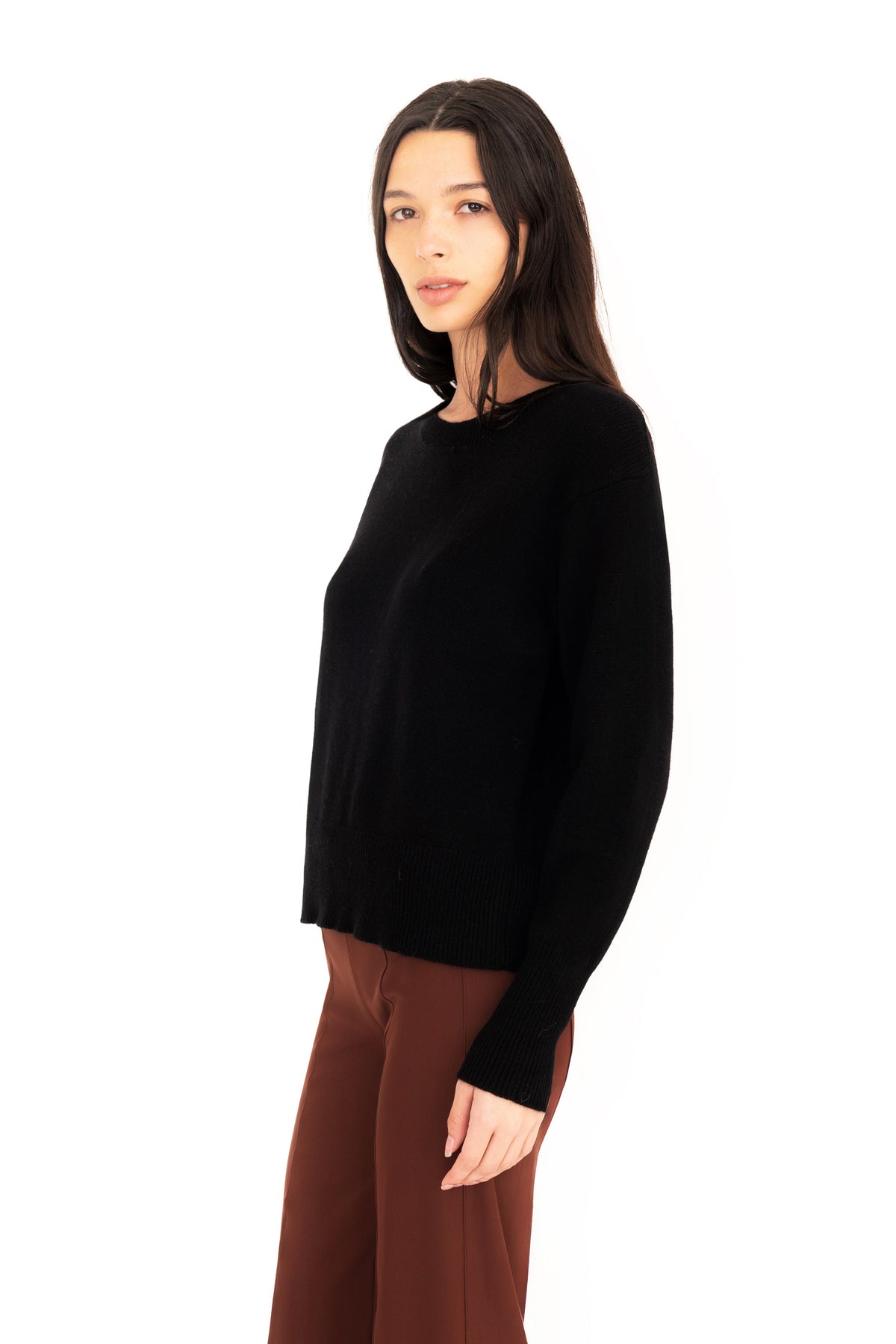 Woman wearing a black crew-neck sweater and dark brown pants on a white studio background.