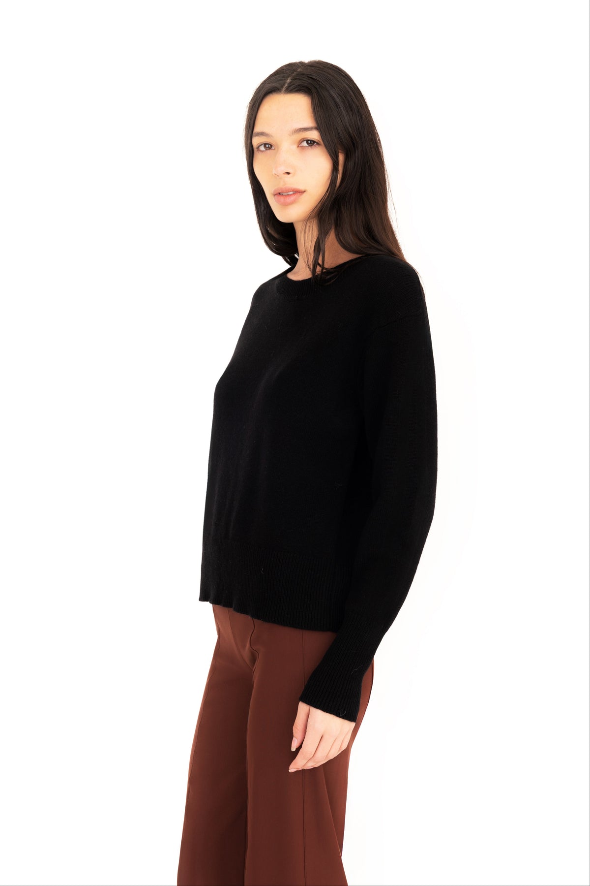 Woman wearing a black crew-neck sweater and dark brown pants on a white studio background.