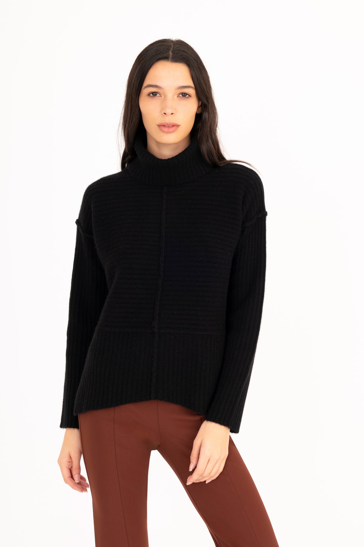 A woman modeling a black ribbed funnel-neck sweater, paired with brown pants, against a plain white studio background.