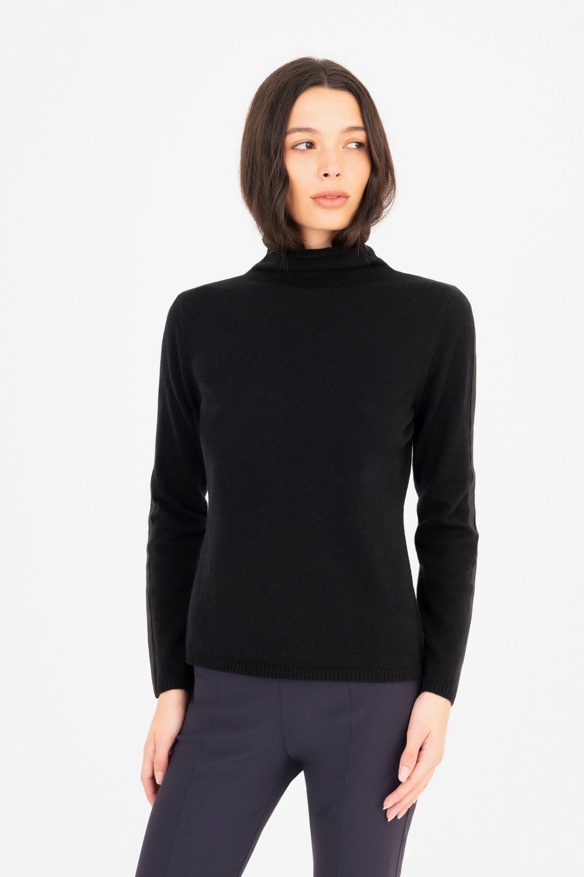 A woman modeling a black long-sleeve mock neck knit sweater, paired with dark navy pants against a plain white studio background.