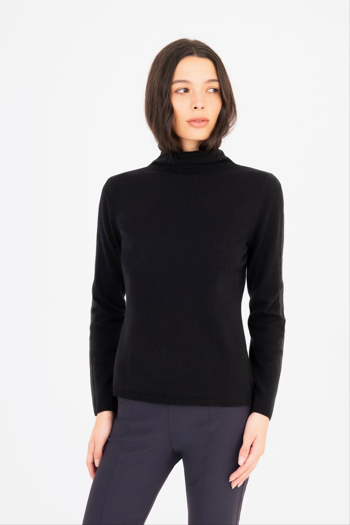 A woman modeling a black long-sleeve mock neck knit sweater, paired with dark navy pants against a plain white studio background.