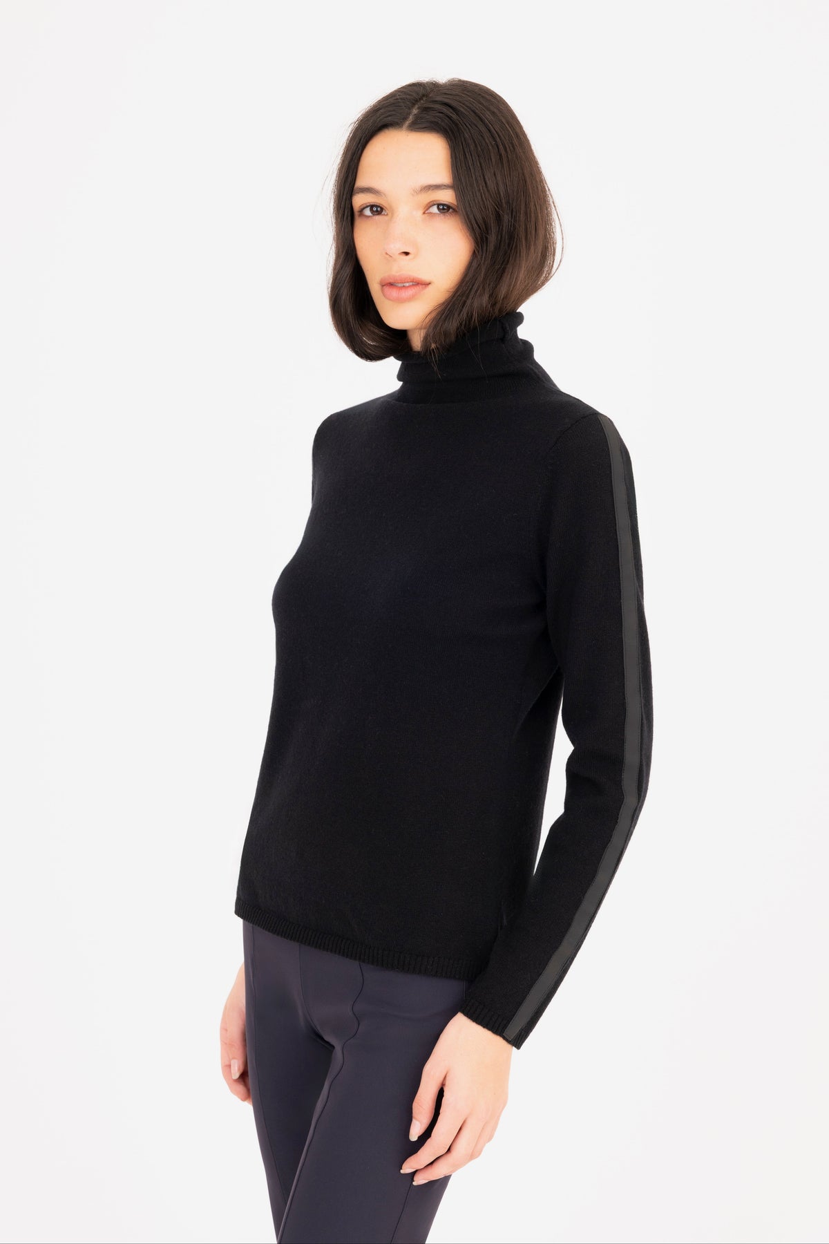 A woman modeling a black turtleneck sweater, paired with dark navy pants, against a plain white studio background.