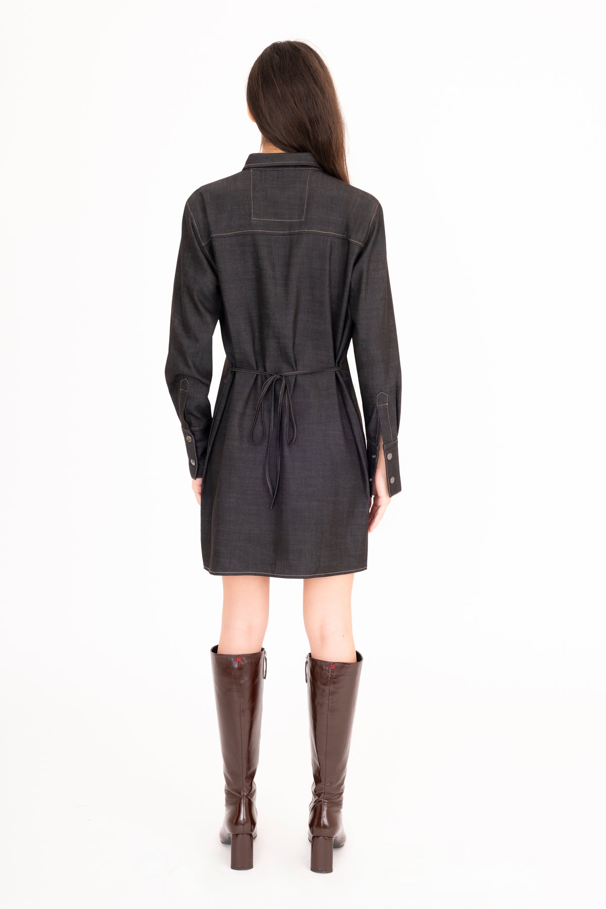 A woman viewed from the back wearing a black denim belted shirt dress and brown knee-high leather boots, standing against a plain white studio background.
