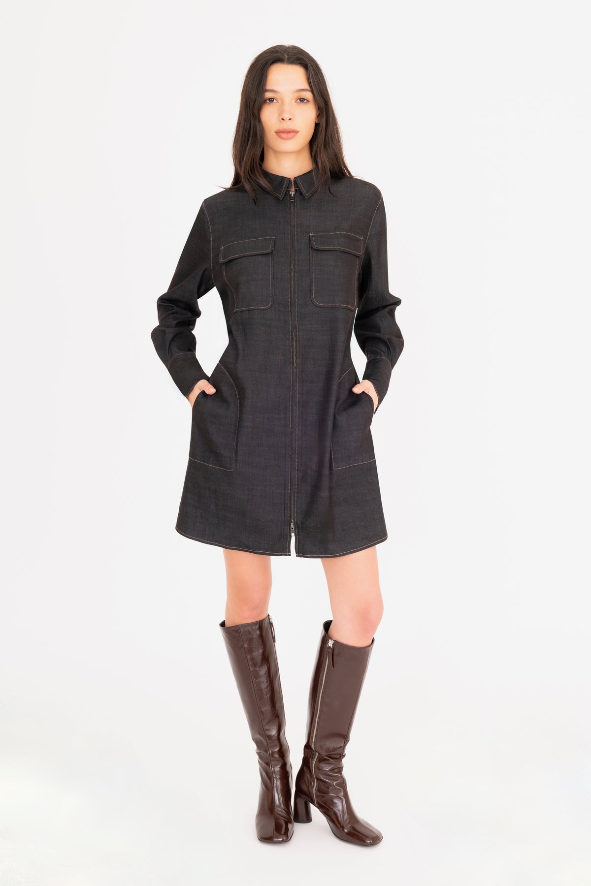 A woman modeling a black denim shirt dress with front pockets, paired with dark brown knee-high leather boots, against a plain white studio background.