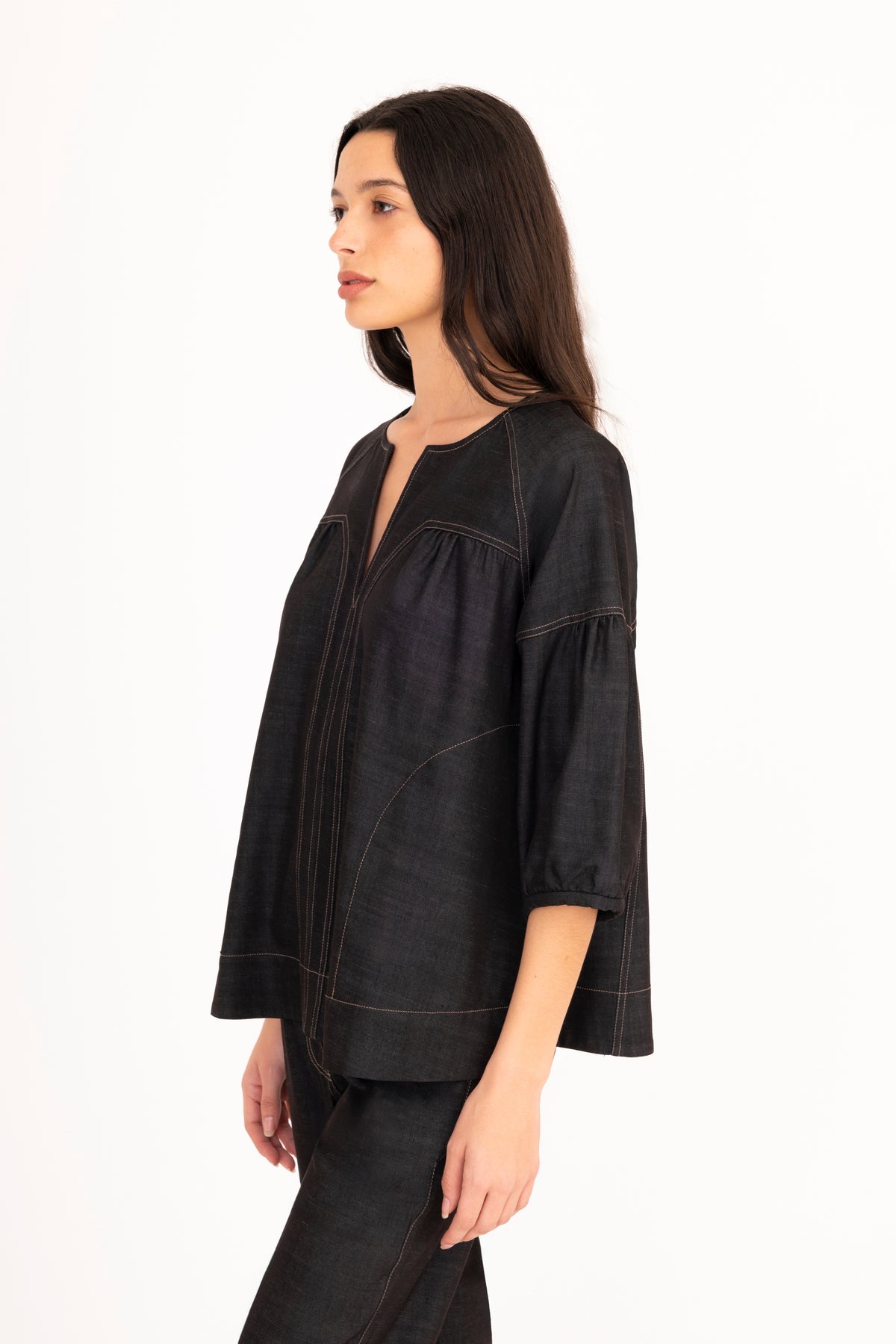 A woman modeling a black denim top with a split neckline, gathered shoulders, and puff sleeves, paired with matching denim pants against a plain white studio background.