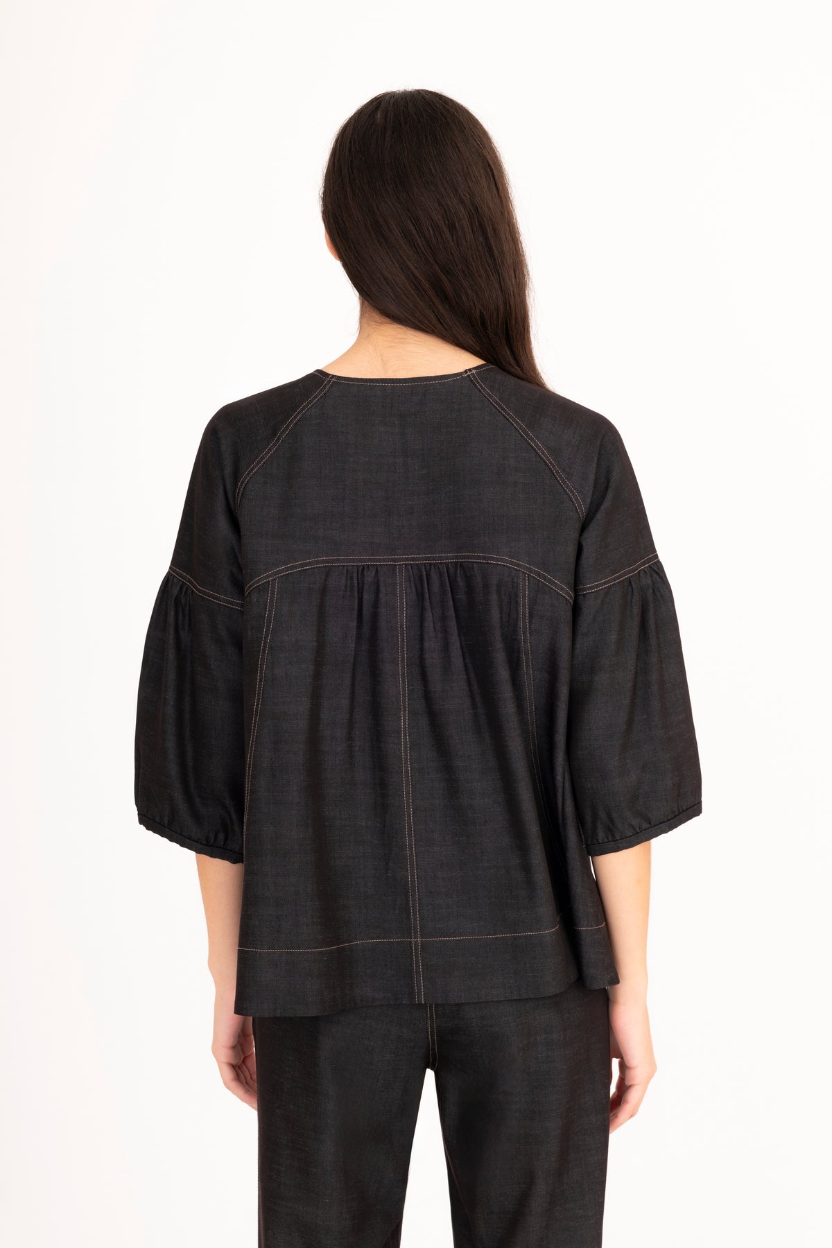 A woman viewed from behind modeling a black denim top with a split neckline, gathered shoulders, and puff sleeves, paired with matching denim pants against a plain white studio background.