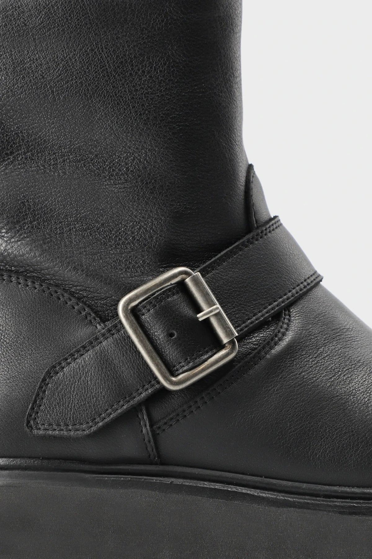 Close-up of a black leather boot featuring a wide strap with a silver-toned rectangular buckle, set against a light gray background.