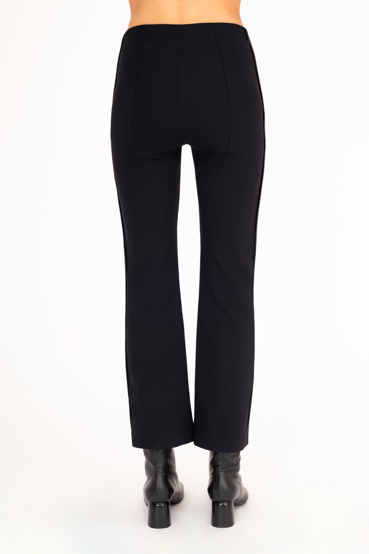 A back view of a model wearing black flared pants with black vegan leather contrast piping and black leather ankle boots, set against a plain white background.
