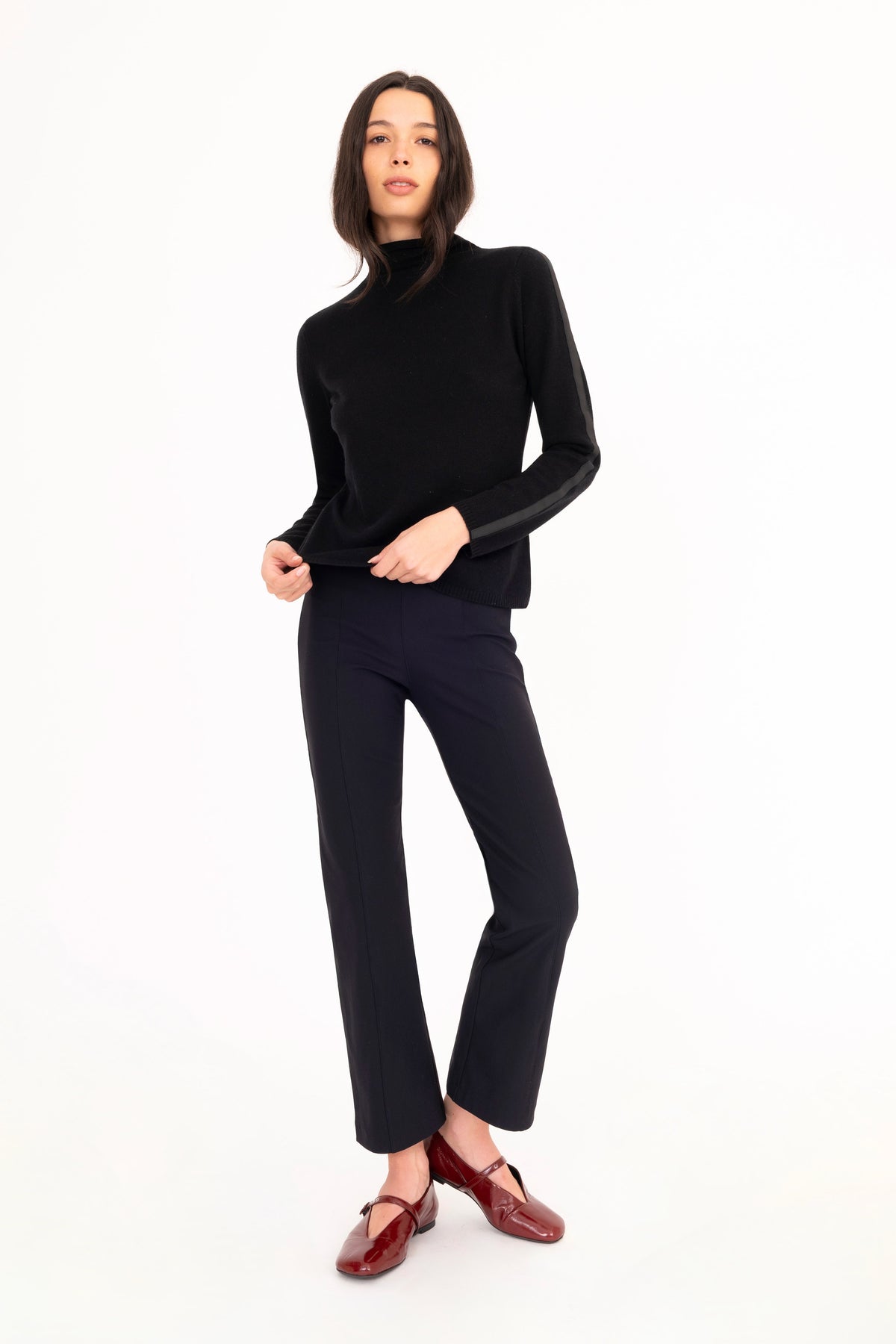 A woman modeling a black long-sleeve mock neck cashmere top and black trousers, paired with dark red mary jane flats, against a solid white studio background.