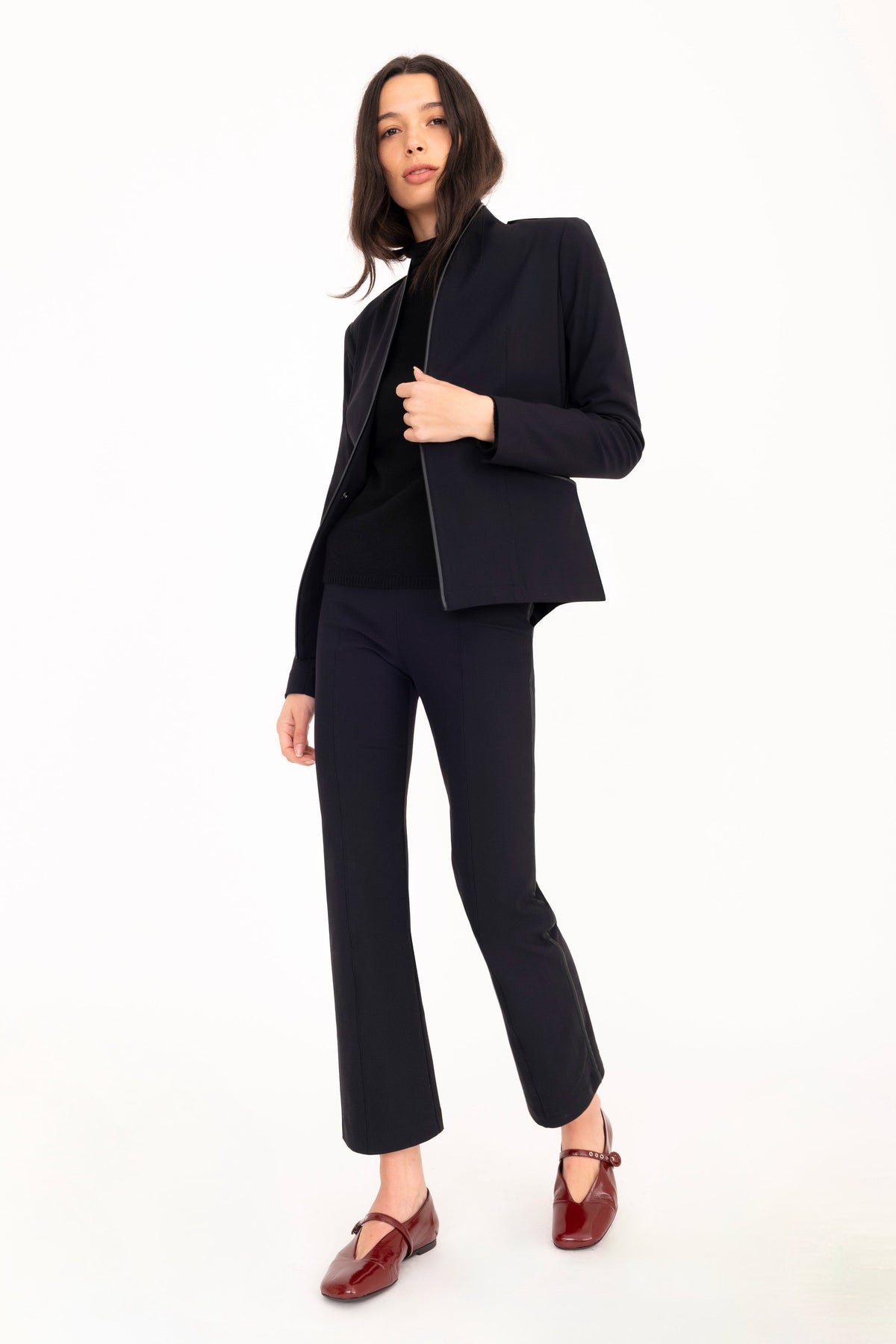 A model poses in a black tailored suit consisting of a blazer and trousers, layered over a black turtleneck sweater and paired with maroon mary jane flats, against a solid white studio background.