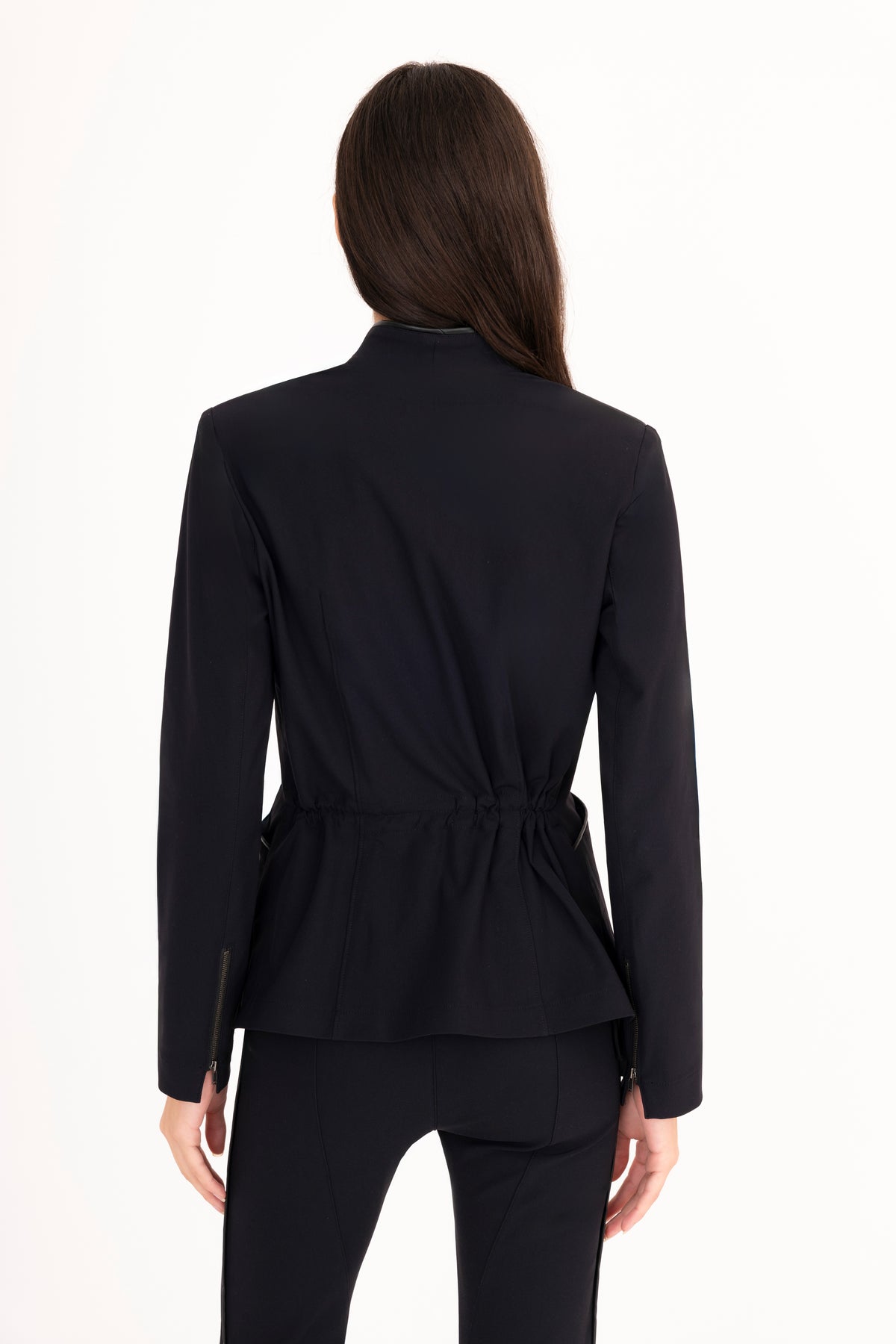 A woman shown from the back wearing a tailored black blazer with a structured collar and peplum waist, paired with black trousers, against a plain white studio background.