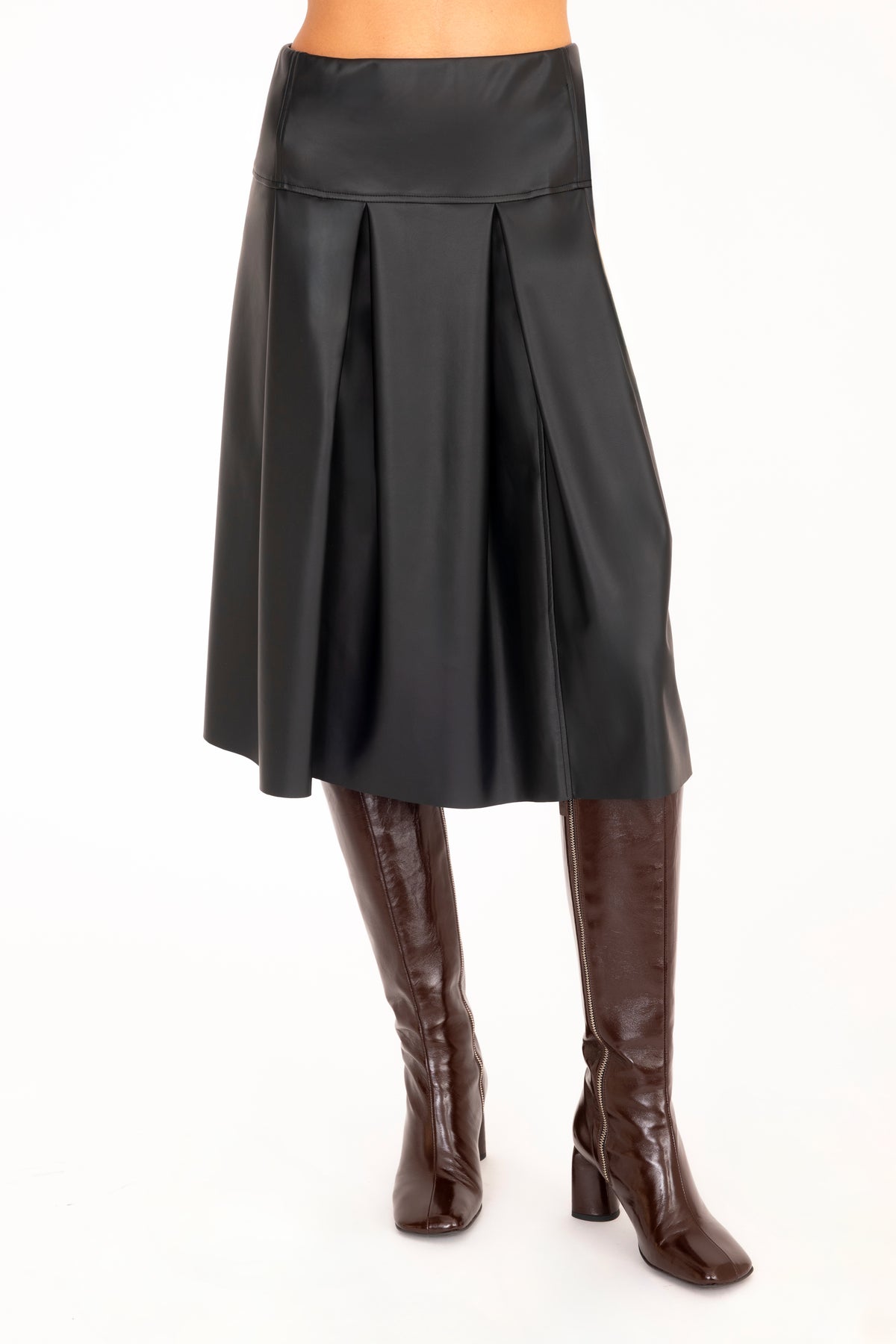 A black vegan leather pleated A-line skirt shown on a model from the waist down, paired with dark brown knee-high leather boots against a plain white background.