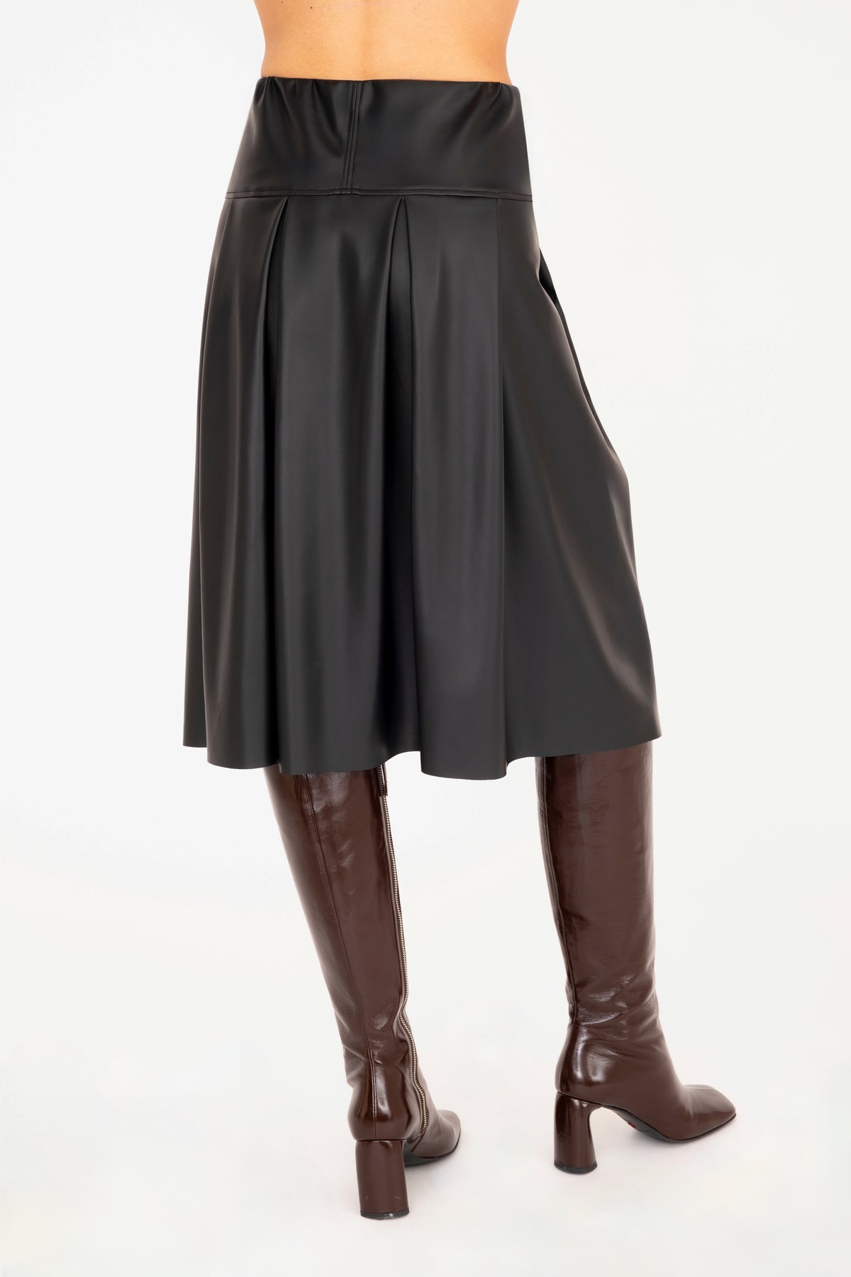 Back view of a black vegan leather pleated A-line skirt shown on a model from the waist down, paired with dark brown knee-high leather boots against a plain white background.
