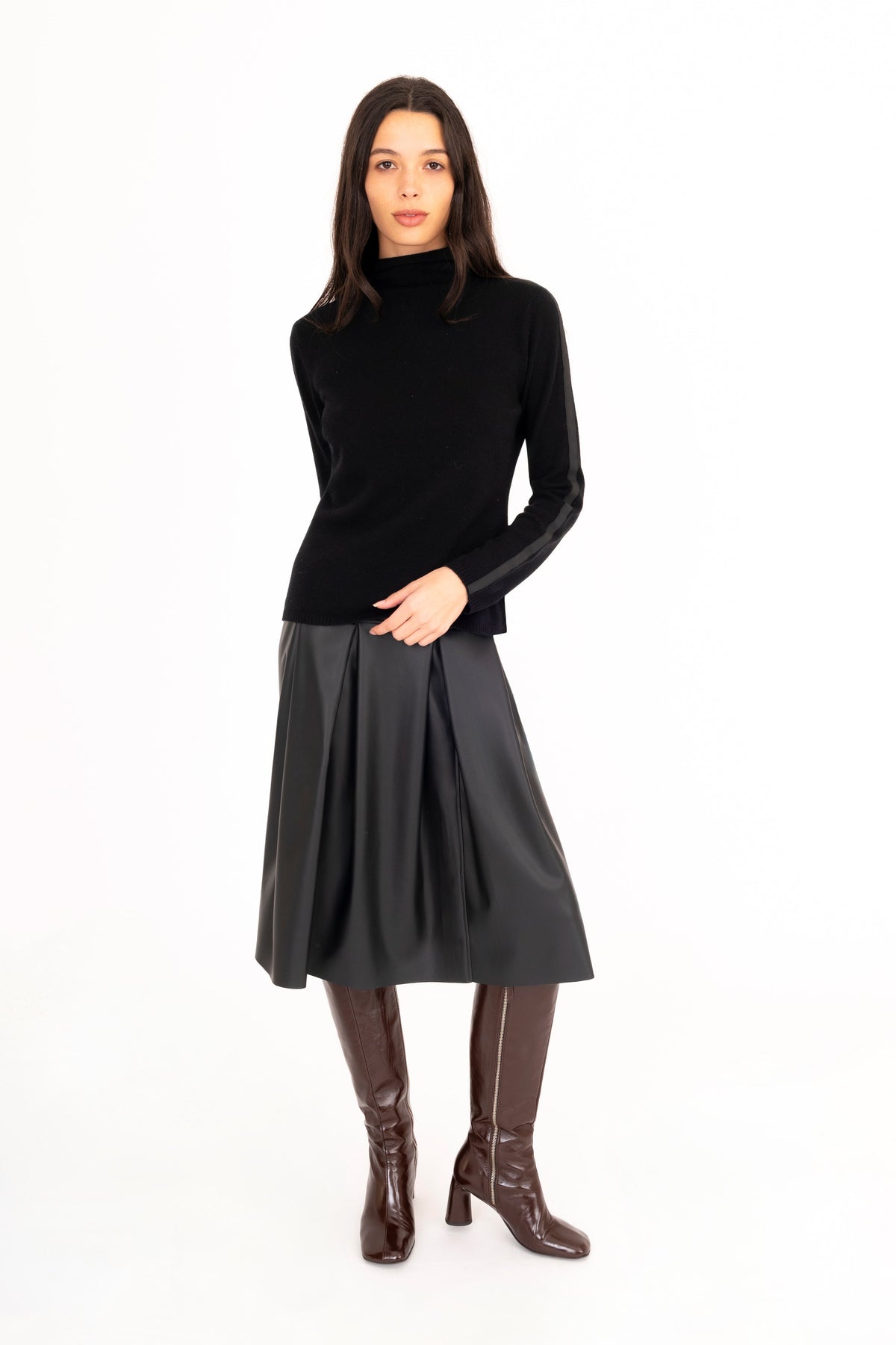 A woman modeling a black long-sleeve cashmere turtleneck sweater, a black vegan leather pleated midi skirt, and dark brown knee-high boots against a plain white studio background.