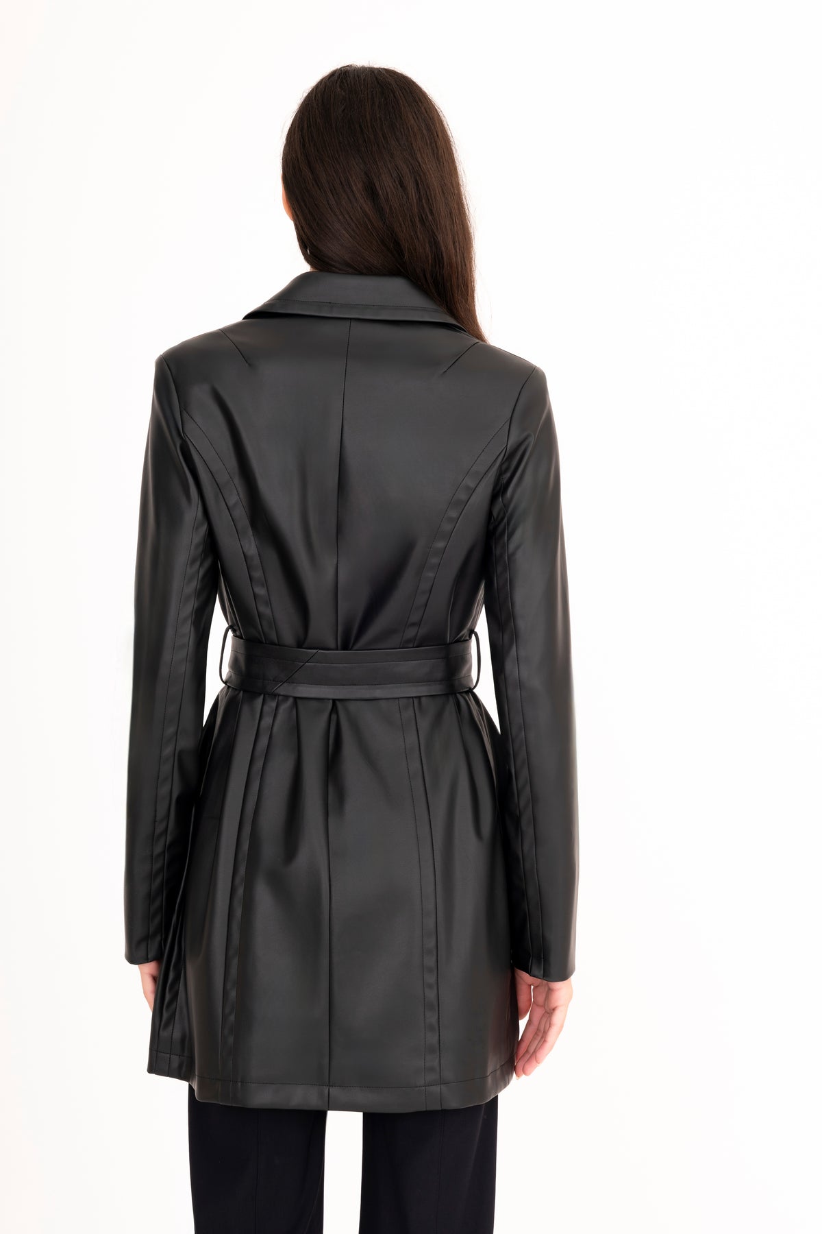 A woman viewed from the back modeling a black vegan leather trench coat with a collar, waist tie, and front pockets, paired with matching black pants, against a plain white studio background.
