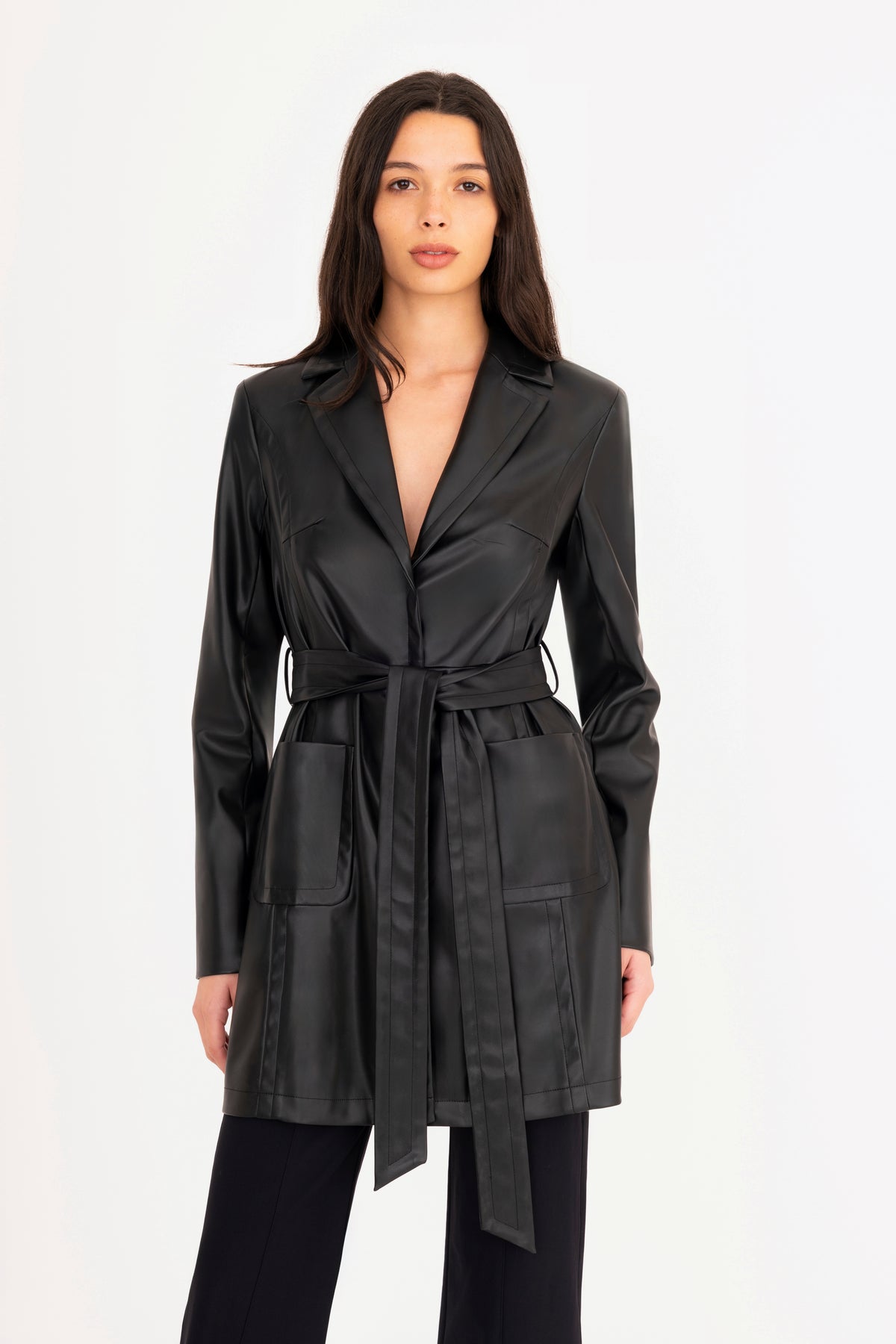 A woman modeling a black vegan leather trench coat with a collar, waist tie, and front pockets, paired with matching black pants, against a plain white studio background.