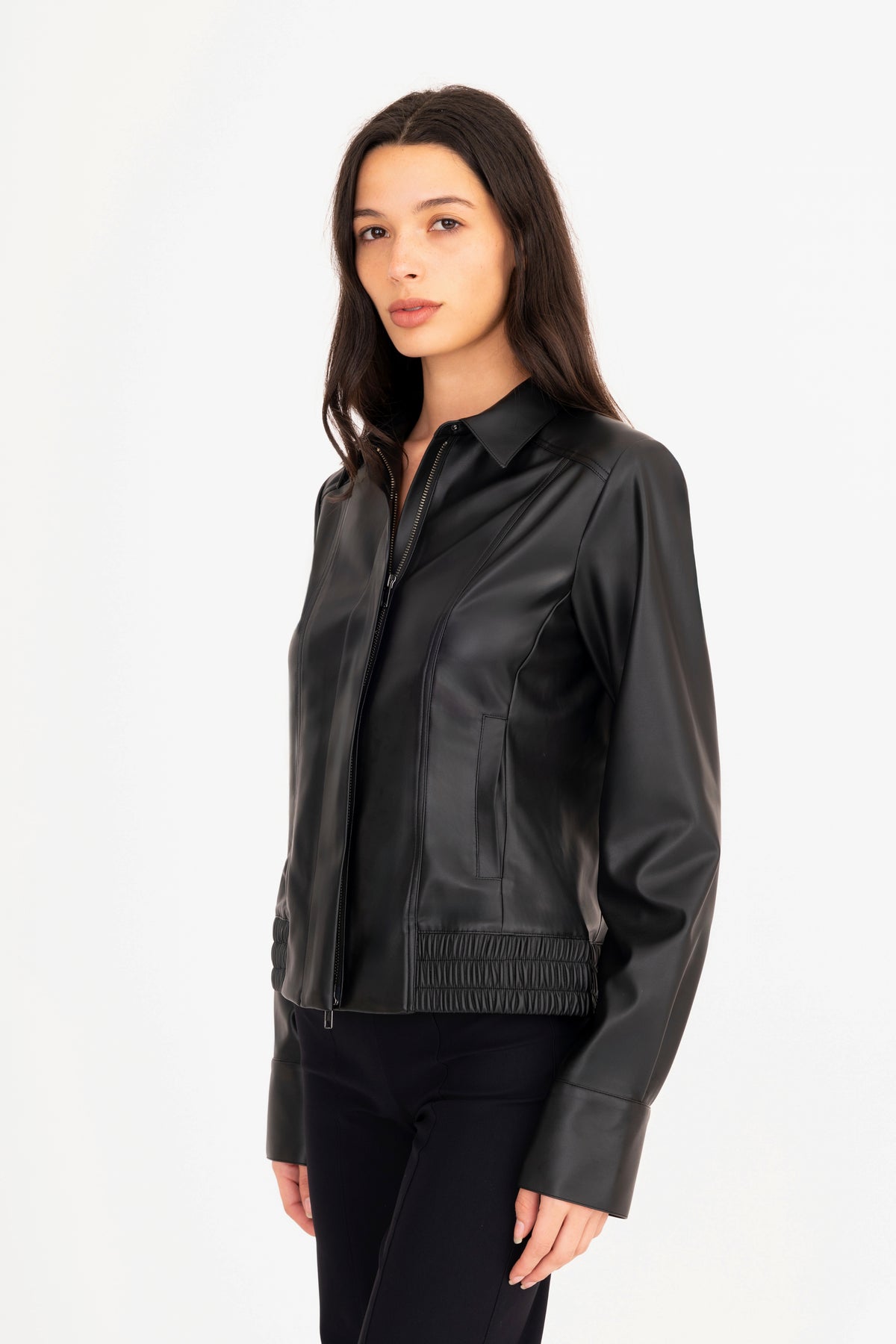 A woman modeling a black vegan leather jacket with a collar, front zipper, and side pockets, paired with matching black pants, against a plain white studio background.