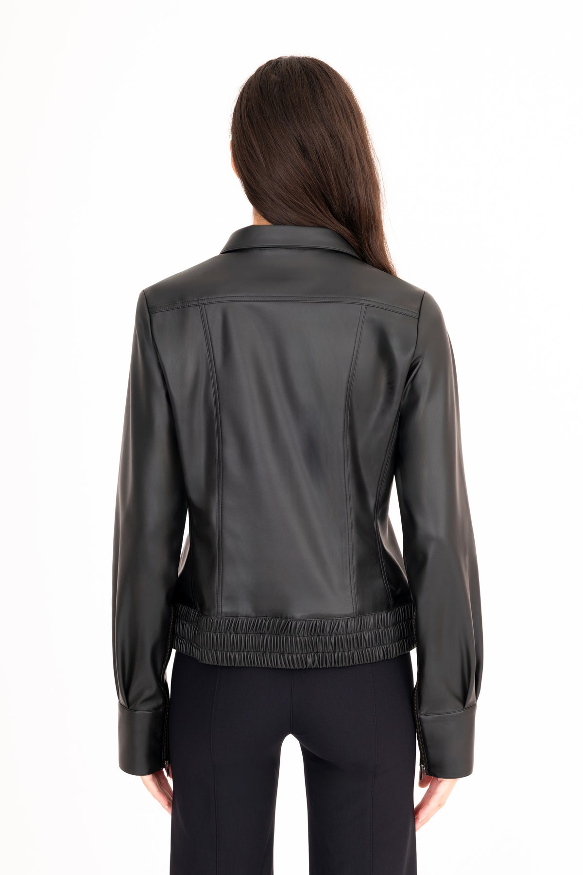 A woman viewed from the back wearing a black vegan leather jacket with a gathered elastic hem, paired with black pants, against a plain white background.