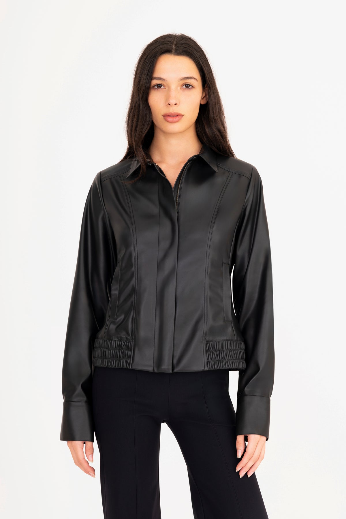 A woman modeling a black vegan leather jacket with a collar, front zipper, and side pockets, paired with matching black pants, against a plain white studio background.