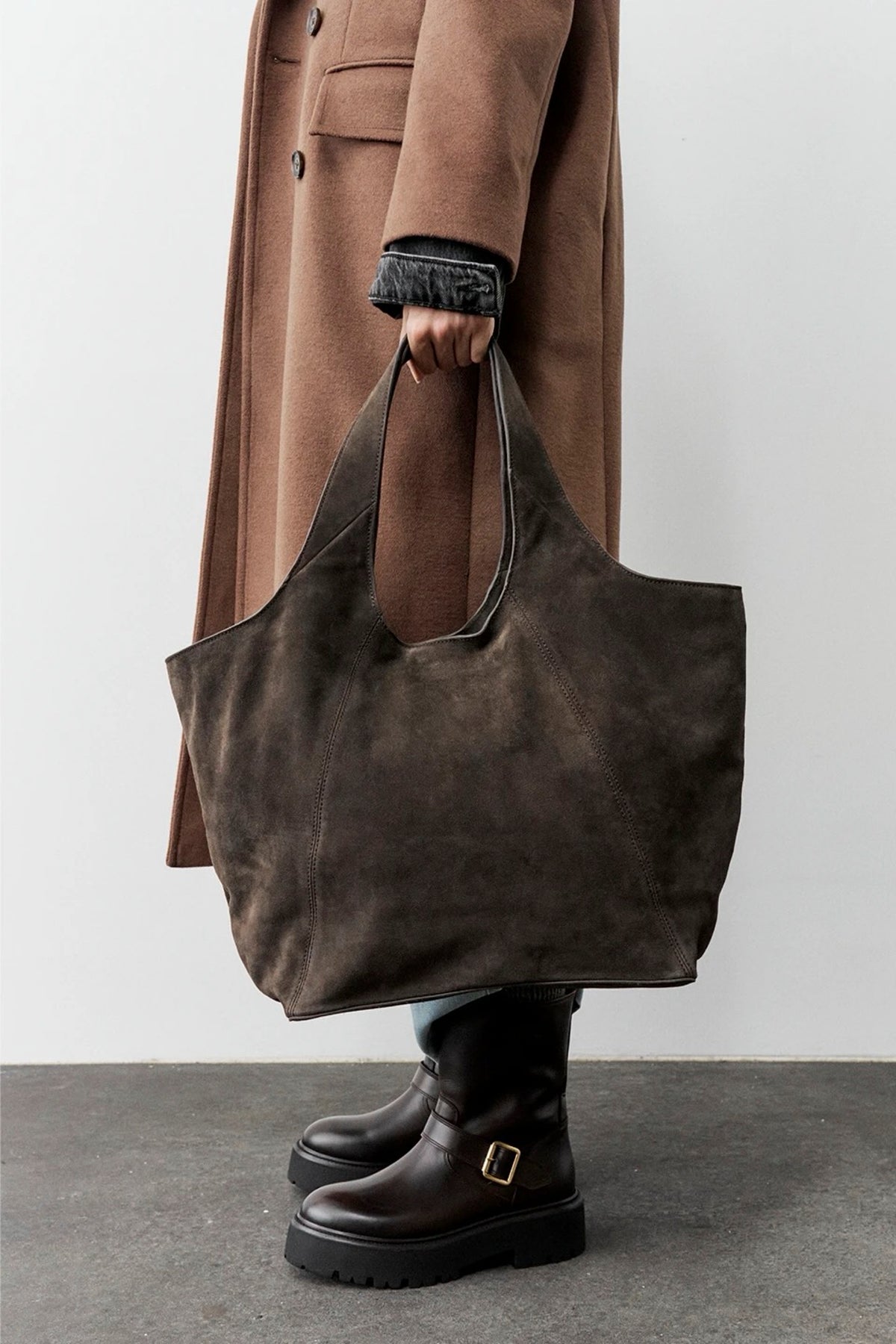 A person wearing a brown coat and dark boots holds a large dark brown suede tote bag against a plain light gray wall and concrete floor.