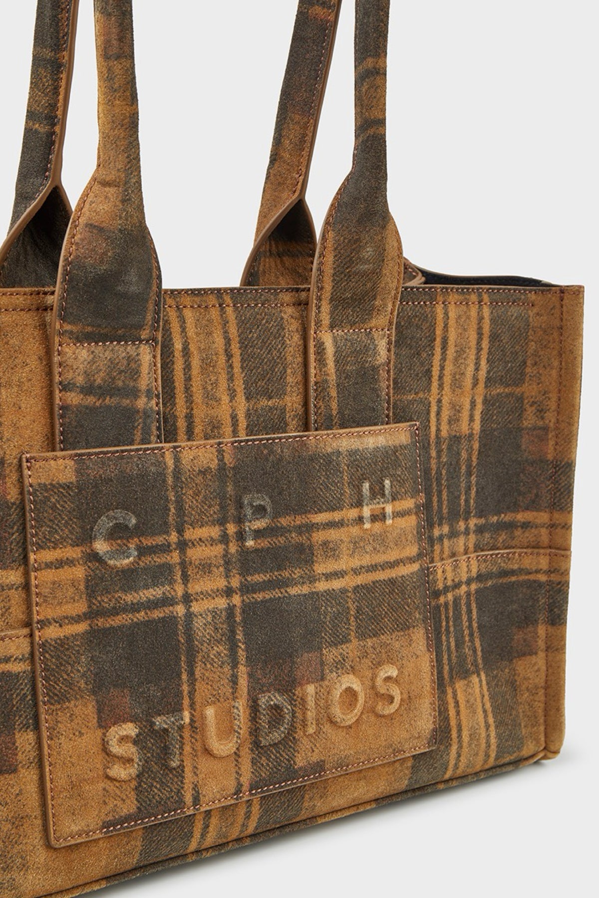 A brown and tan plaid suede tote bag with embossed 'CPH STUDIOS' branding on the front pocket, shown against a light gray background.