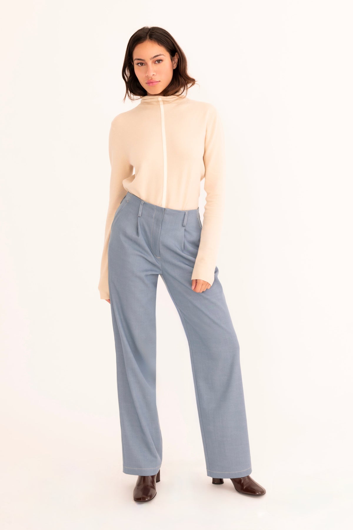 A woman stands against a plain white studio background wearing a light beige mock-neck knit top and high-waisted, wide-leg blue trousers.
