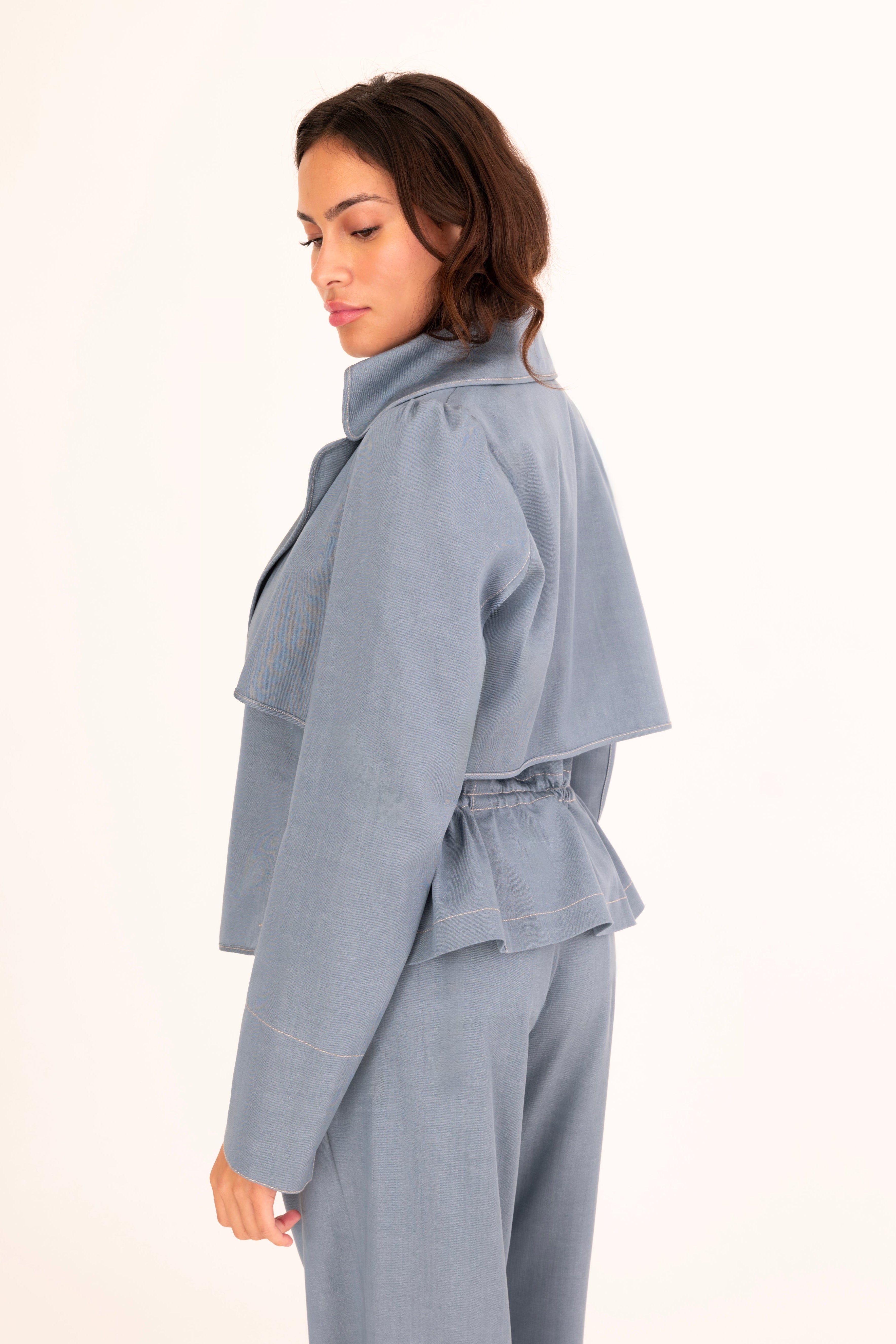 A woman modeling a light blue tailored denim suit with structured shoulders, flap pockets, and matching trousers, worn over a cream mock-neck top against a plain off-white studio background.