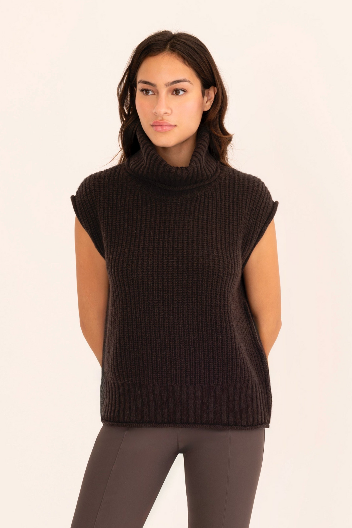 A woman modeling a dark brown ribbed knit sleeveless turtleneck sweater vest against a plain off-white background.
