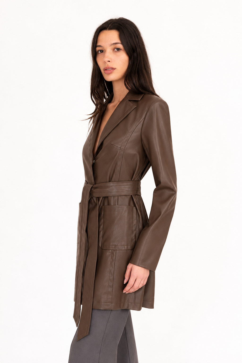 A woman modeling a dark brown vegan leather trench coat with a collar, waist tie, and front pockets, paired with matching dark brown pants, against a plain white studio background.