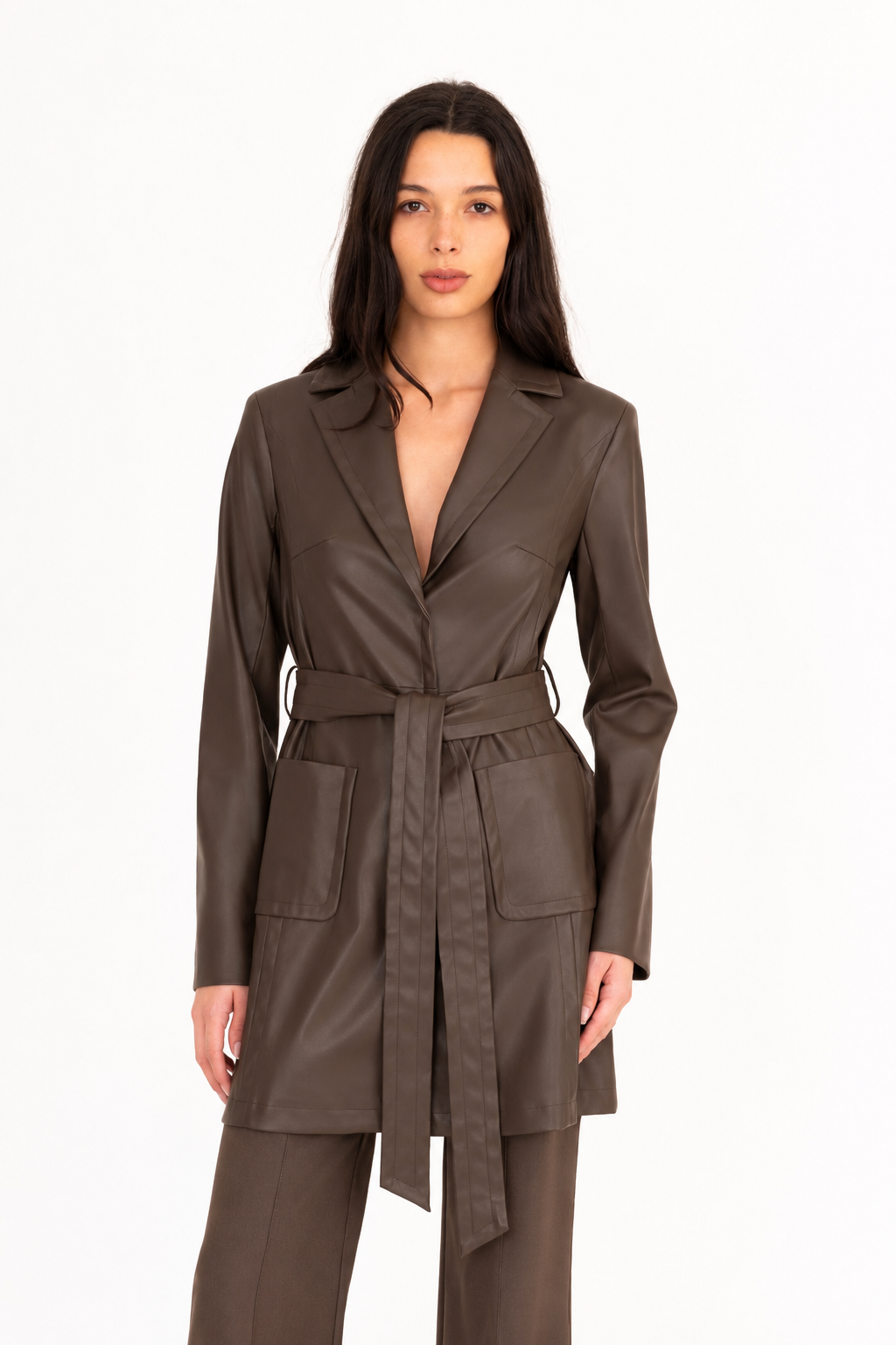A woman modeling a dark brown vegan leather trench coat with a collar, waist tie, and front pockets, paired with matching dark brown pants, against a plain white studio background.
