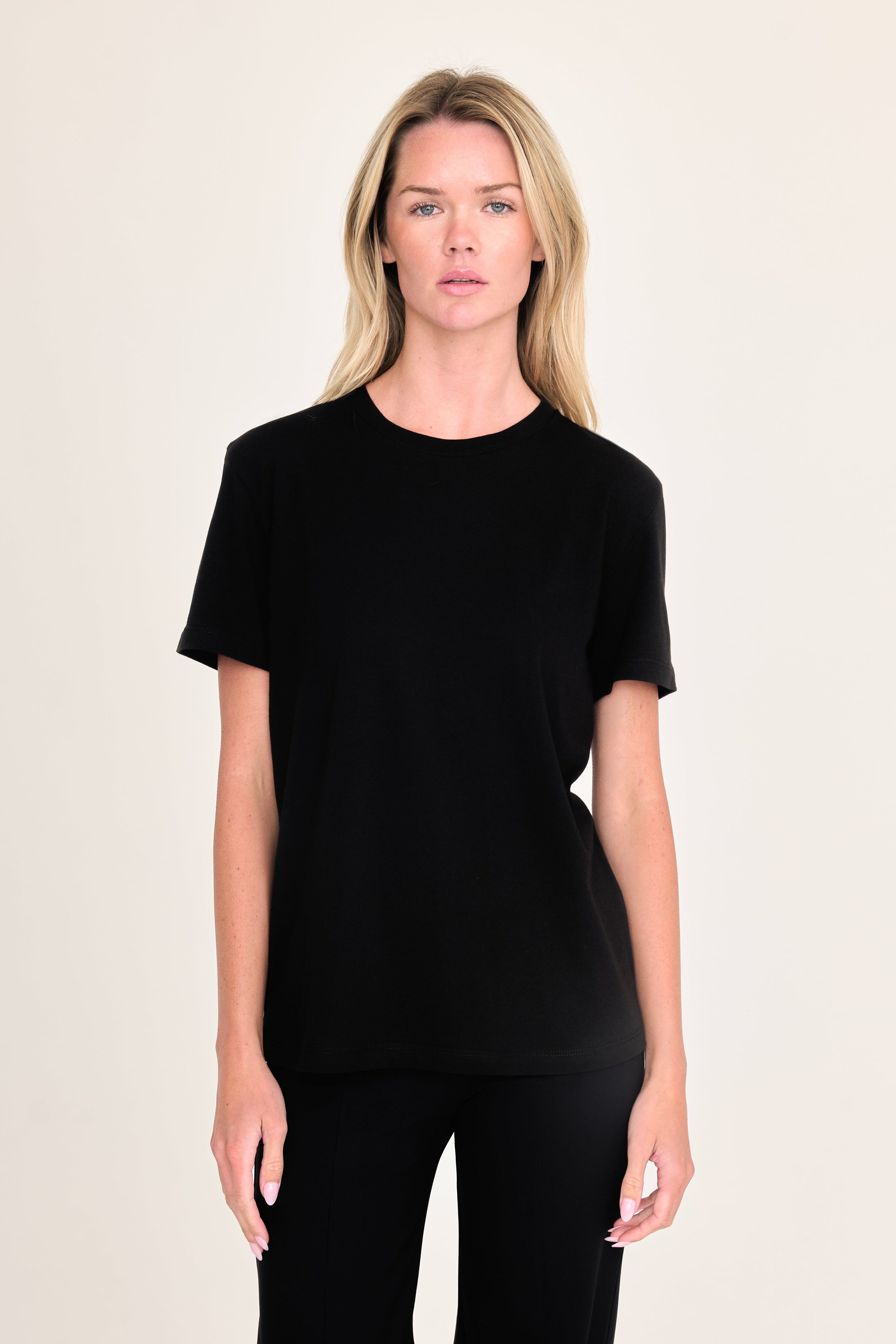 A blonde woman modeling a black short-sleeve crewneck t-shirt and black pants against a solid light beige background.