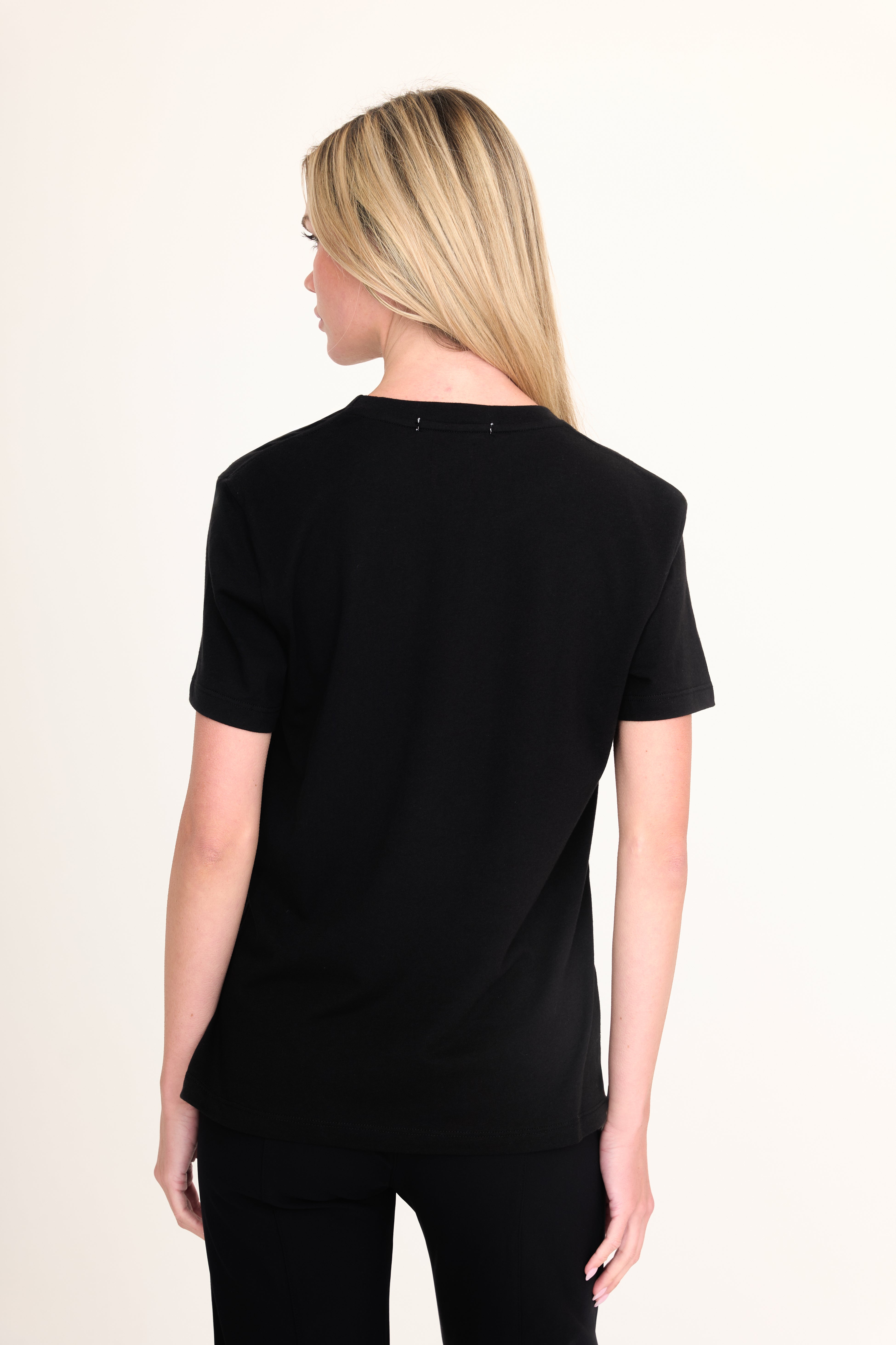 A woman with blonde hair is shown from the back wearing a black short-sleeve crewneck t-shirt and black pants against a plain off-white background.