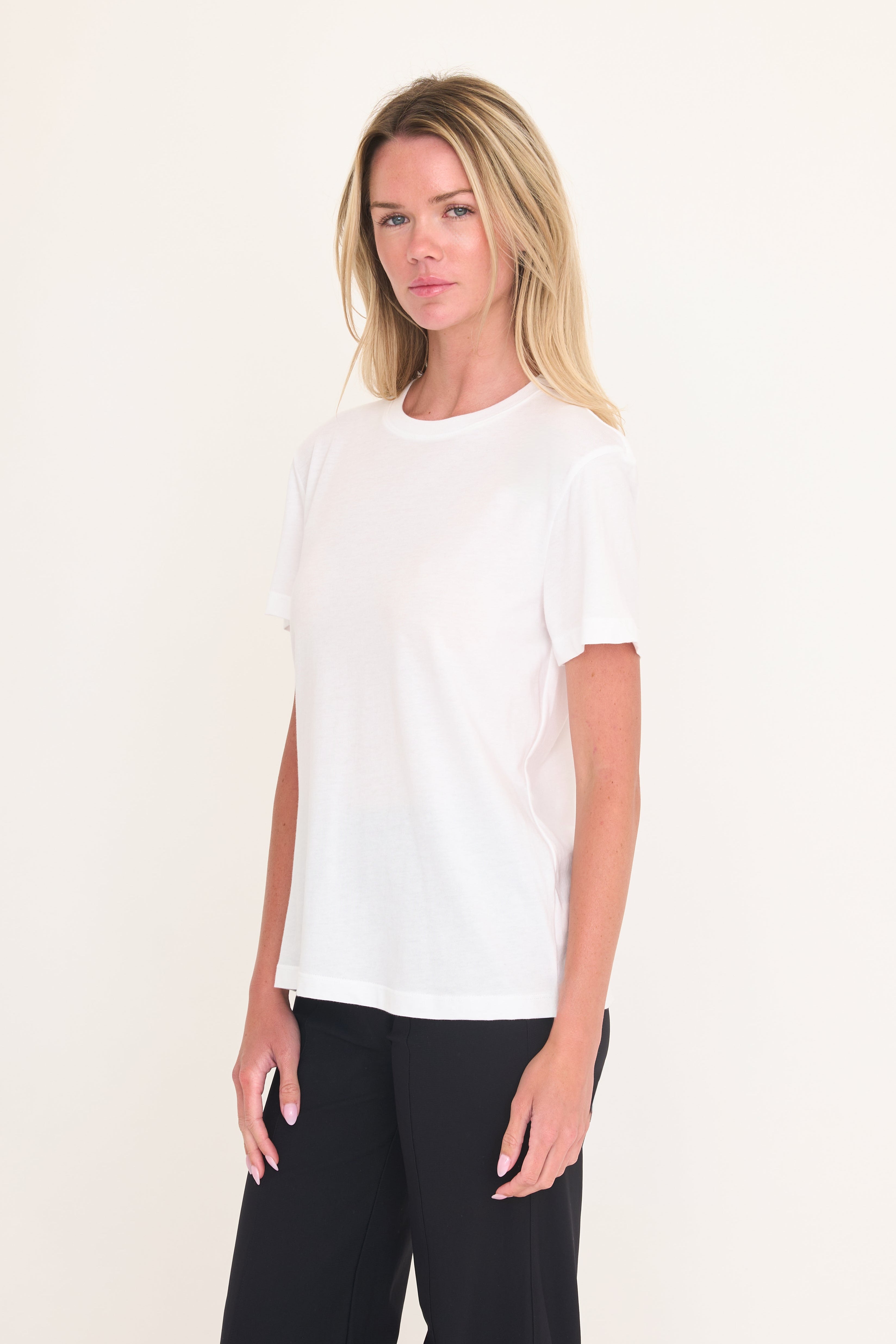 A blonde woman modeling a plain white short-sleeve crewneck t-shirt paired with black pants, standing against a solid off-white studio background.
