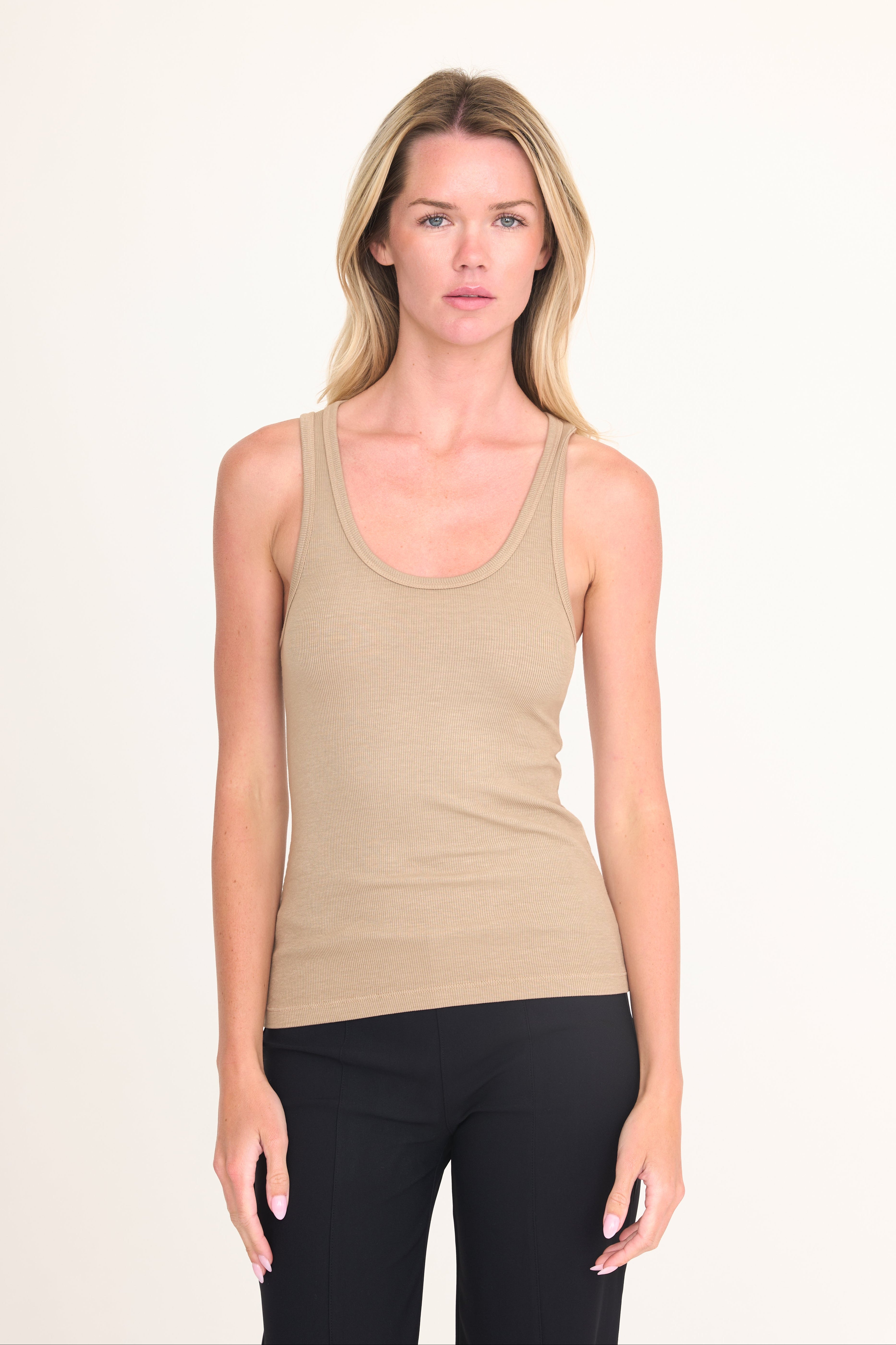 A woman modeling a beige ribbed scoop-neck tank top paired with black pants, standing against a plain off-white studio background.