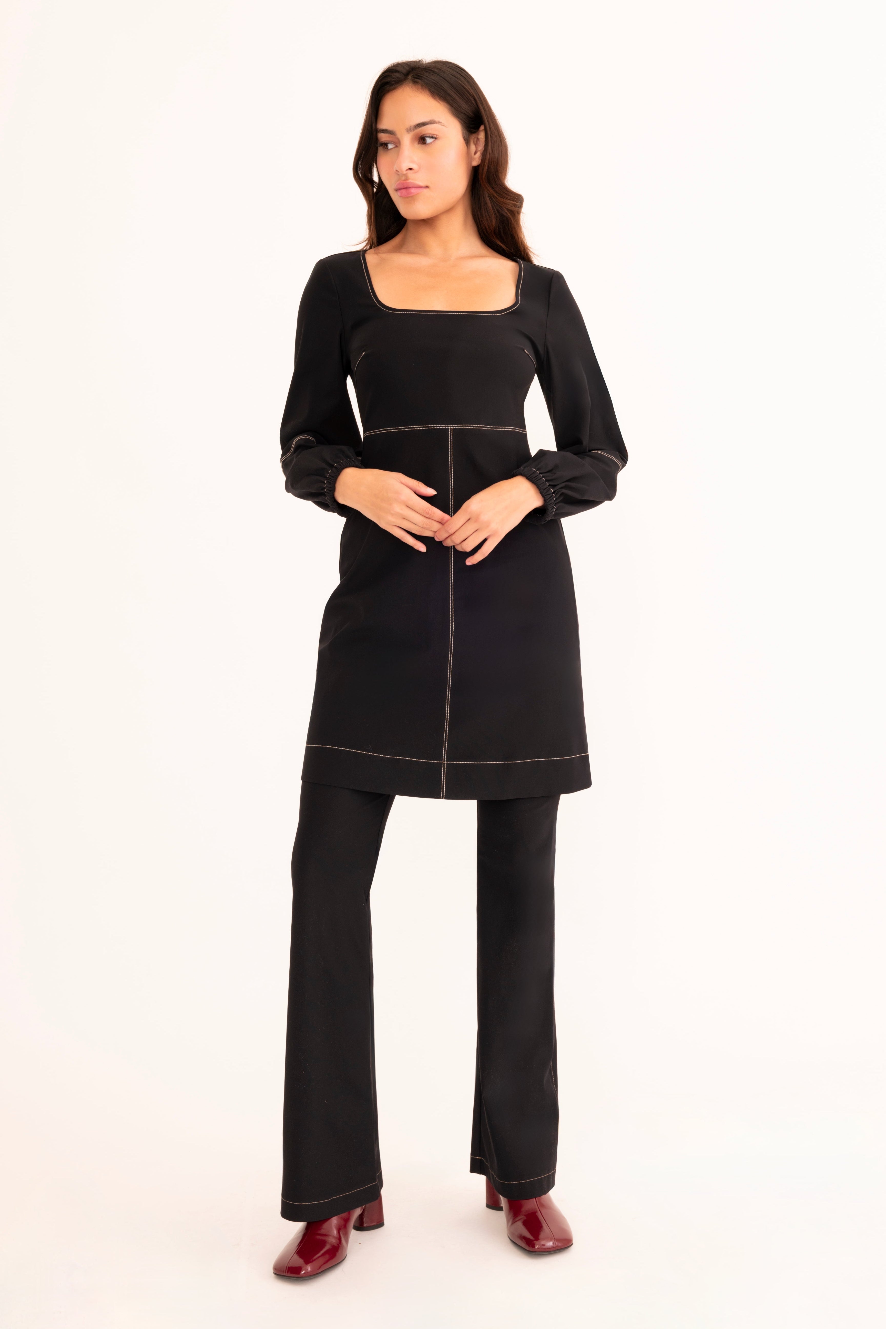 A woman modeling a black long-sleeve tunic dress with a square neckline, gathered waist, and matching black wide-leg pants against a plain white studio background.