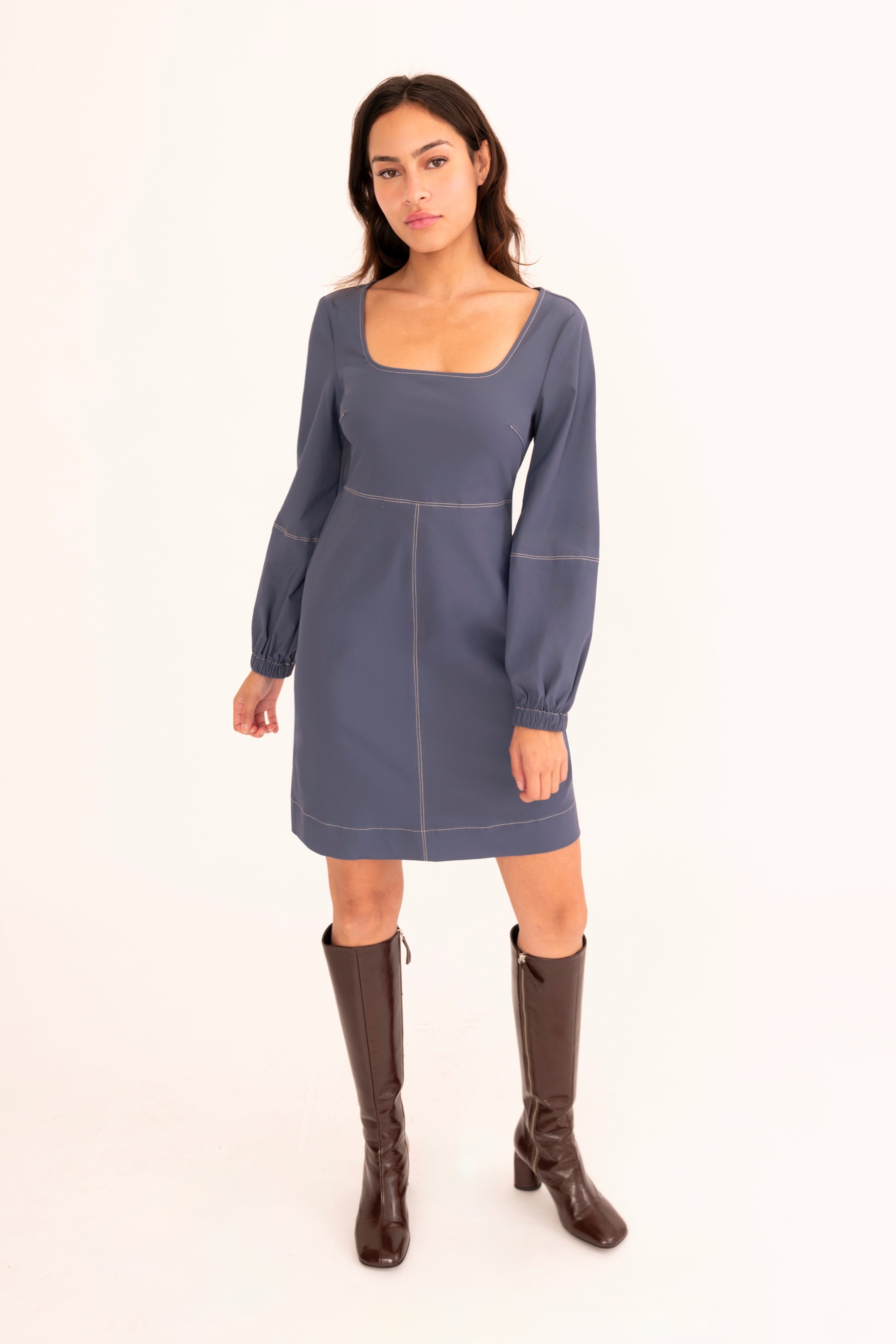 A woman modeling a slate blue long-sleeve mini dress with a square neckline and brown knee-high boots, standing against a plain white studio background.